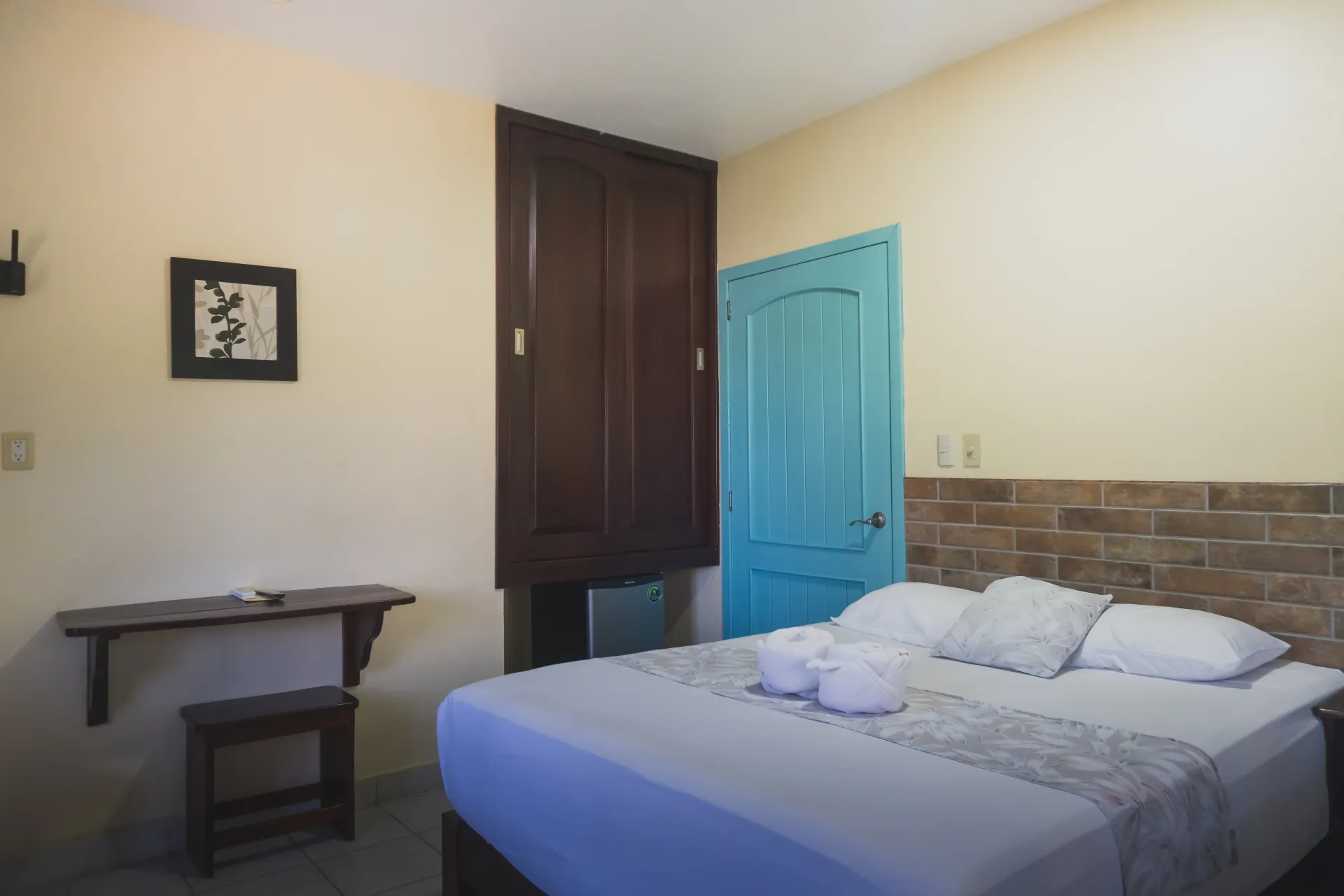 Habitación pequeña con cama, puerta turquesa, armario oscuro, estante de pared y cuadro en la pared.