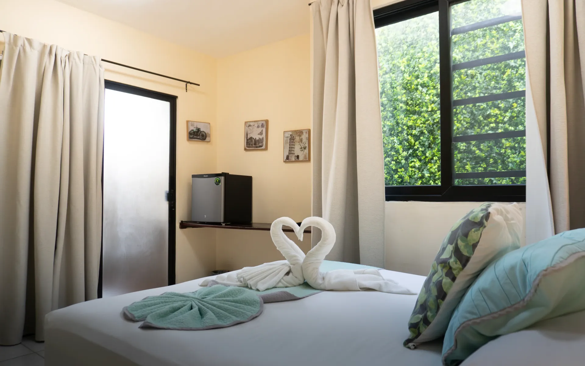 Habitación luminosa con una cama bien hecha, cisnes de toalla y luz solar que entra por las cortinas y una ventana.