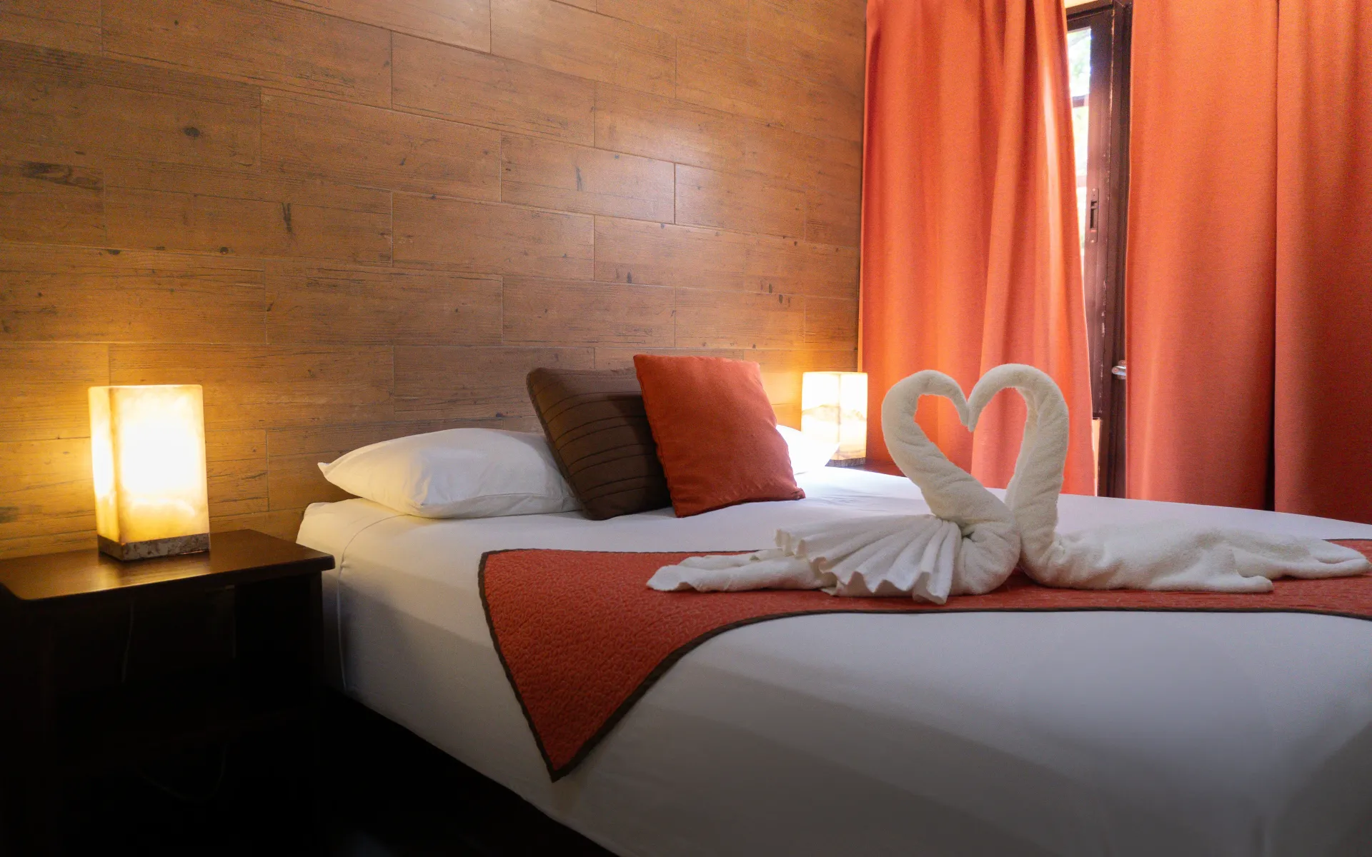 Una acogedora cama de hotel con toallas blancas dobladas en forma de corazón, detalles en naranja.