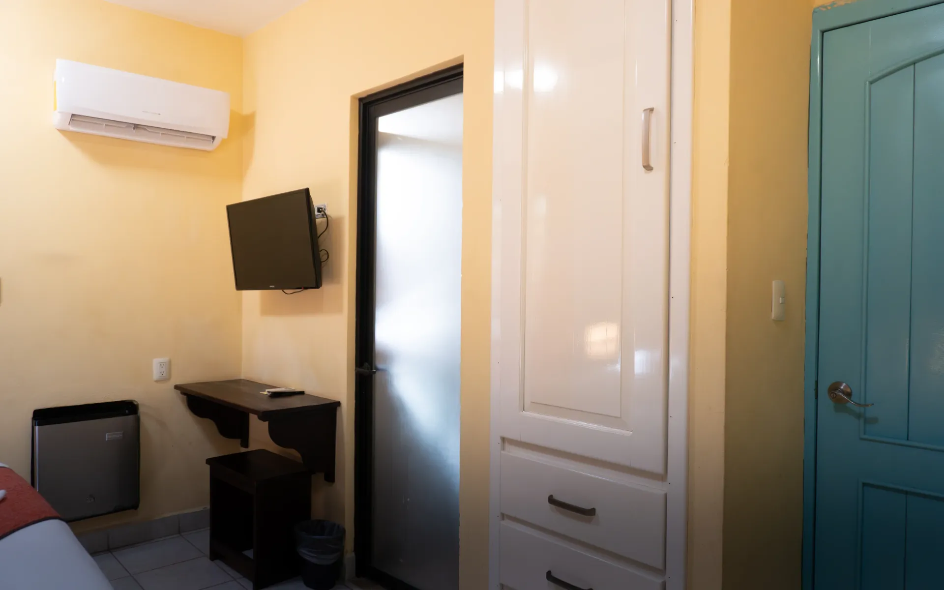 Habitación pequeña con televisor montado en la pared, aire acondicionado, escritorio, armario y puerta color turquesa.