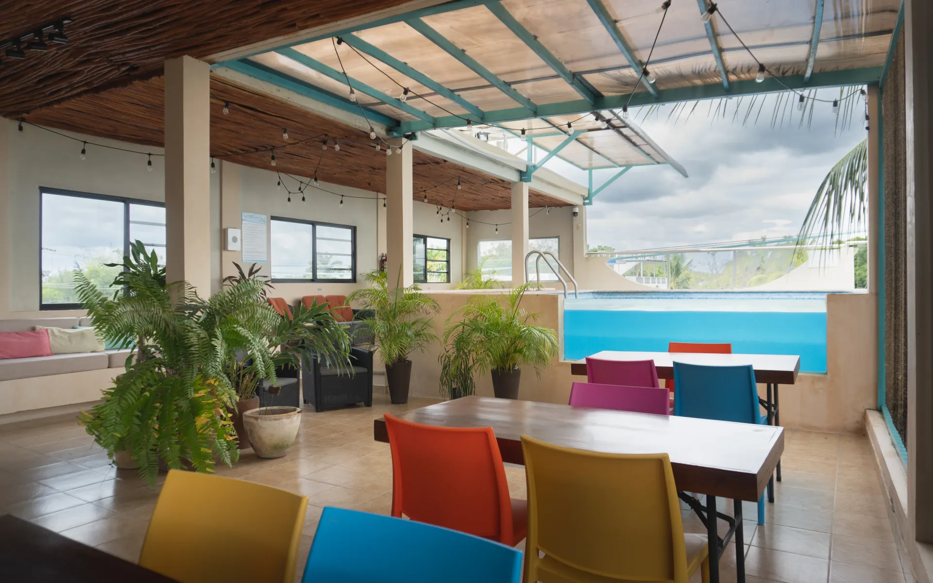 Terraza cubierta en la azotea con sillas y mesas de colores, plantas en macetas y vistas a una piscina de color.