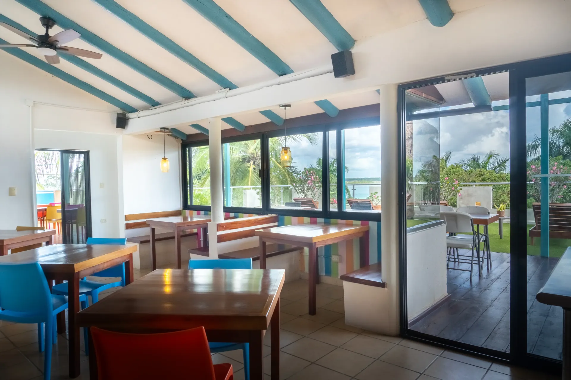 Zona de cafetería luminosa con vigas azules en el techo, mesas de madera y grandes ventanales con vistas a la vegetación.