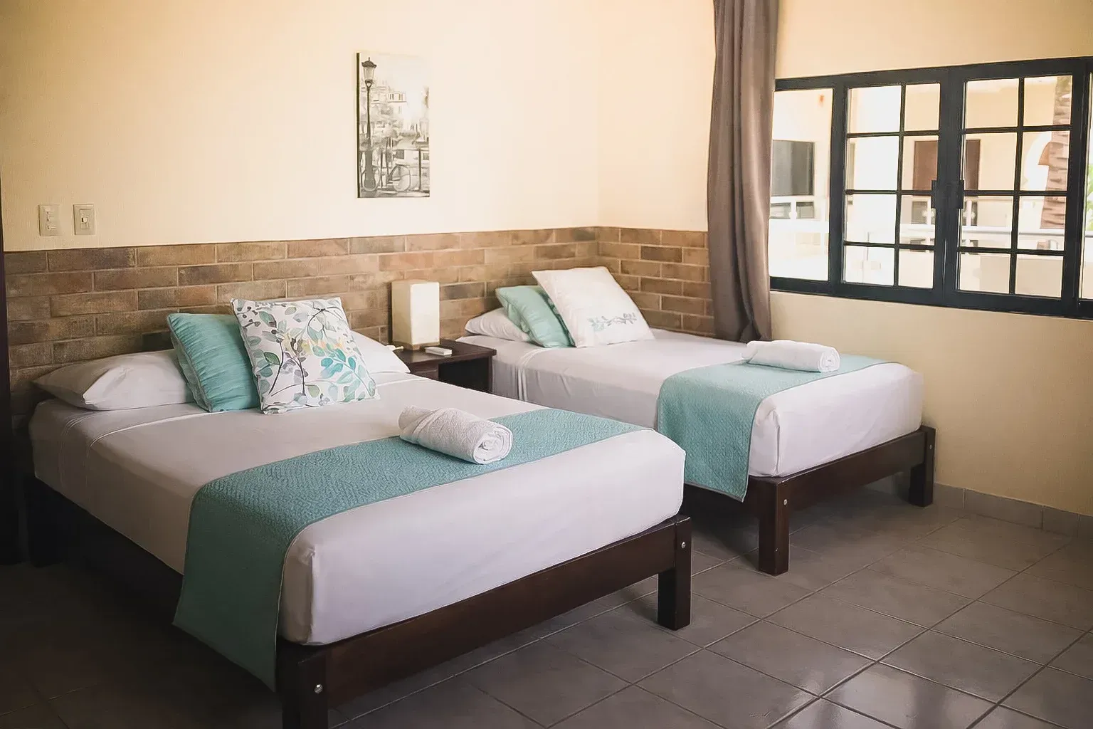 Dos camas individuales con detalles en color turquesa en una habitación luminosa junto a una ventana.