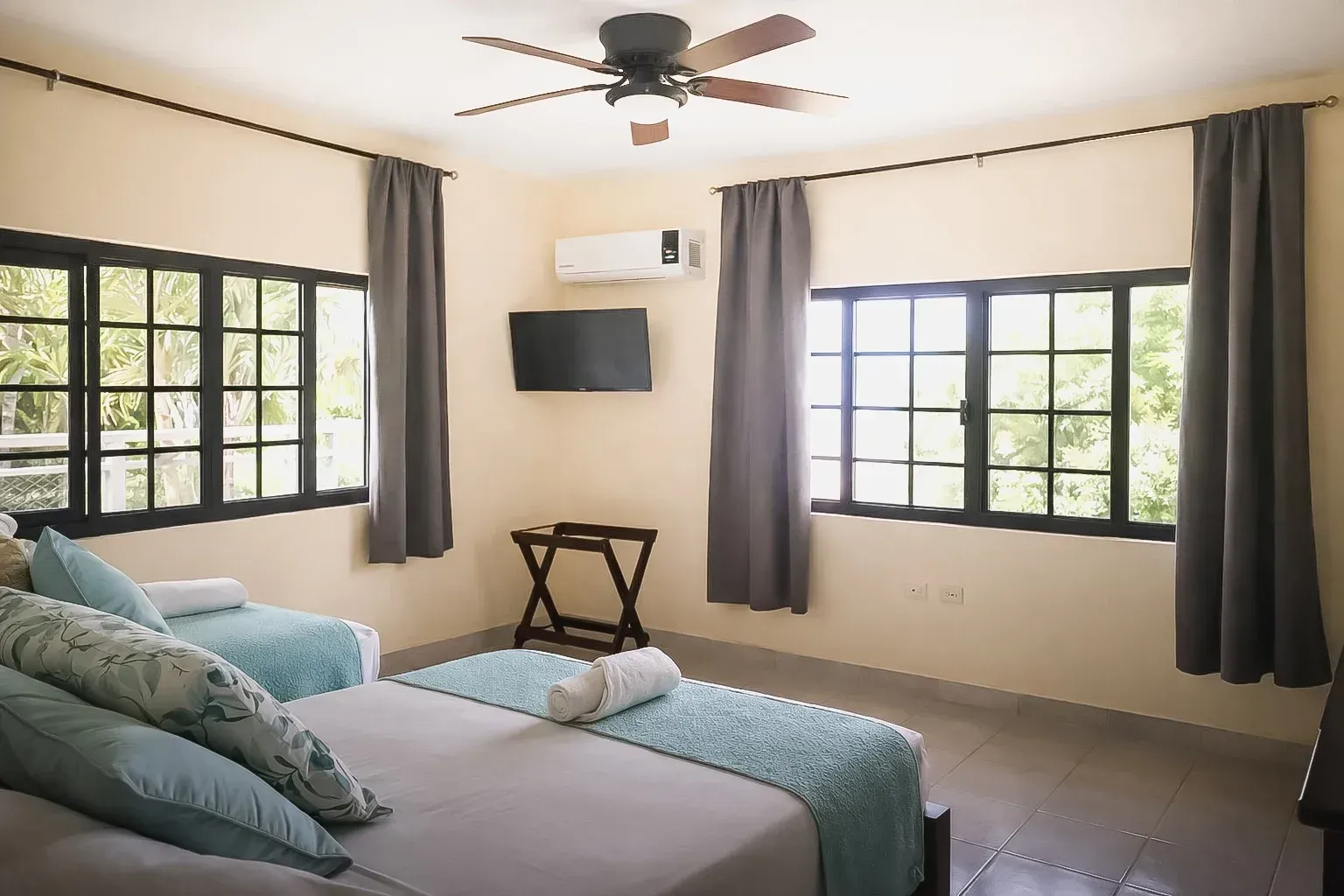 Habitación luminosa con dos ventanas, cama color turquesa, televisor montado en la pared y ventilador.