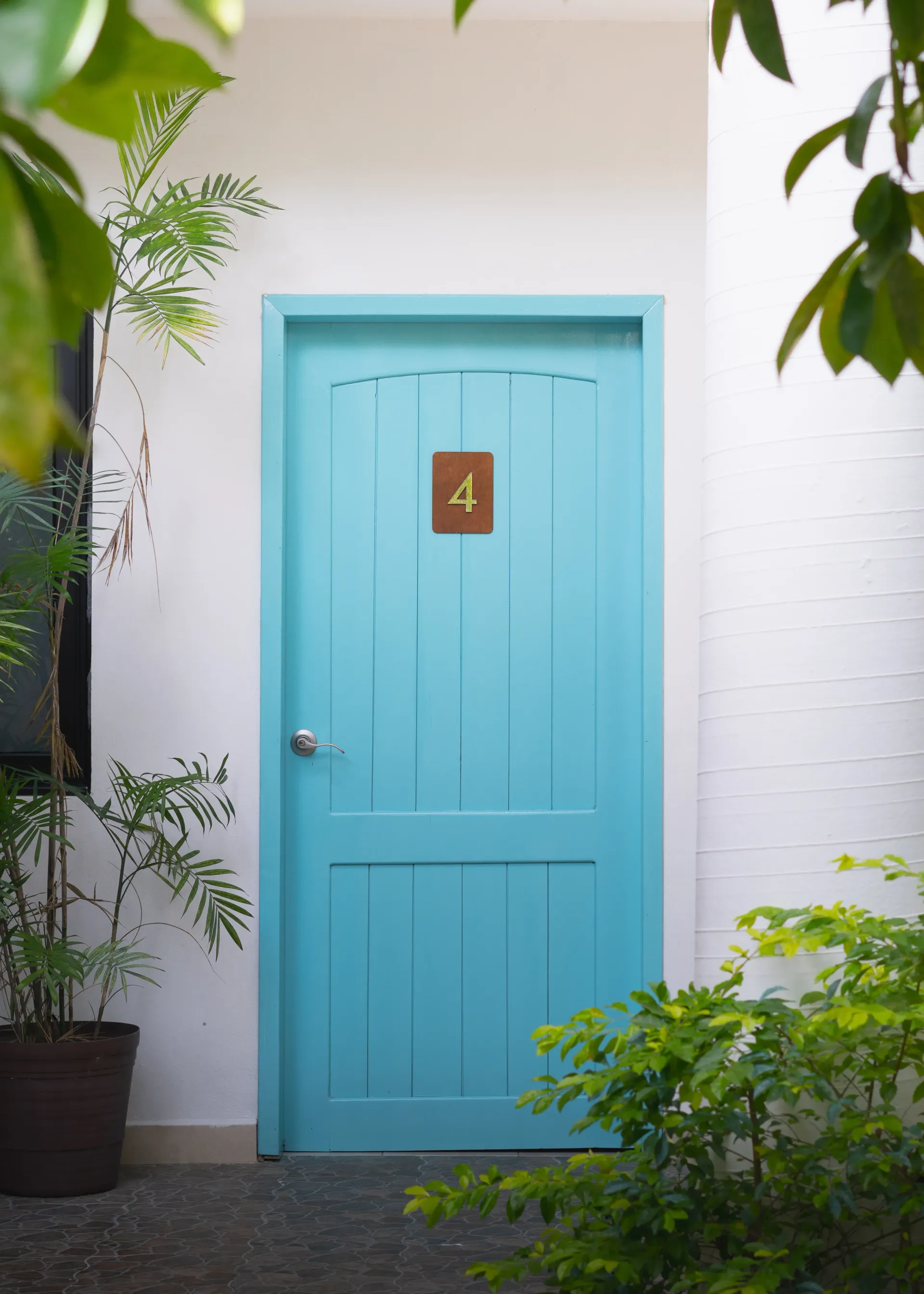 Puerta turquesa con una pequeña placa numerada, enmarcada por paredes blancas y vegetación frondosa.