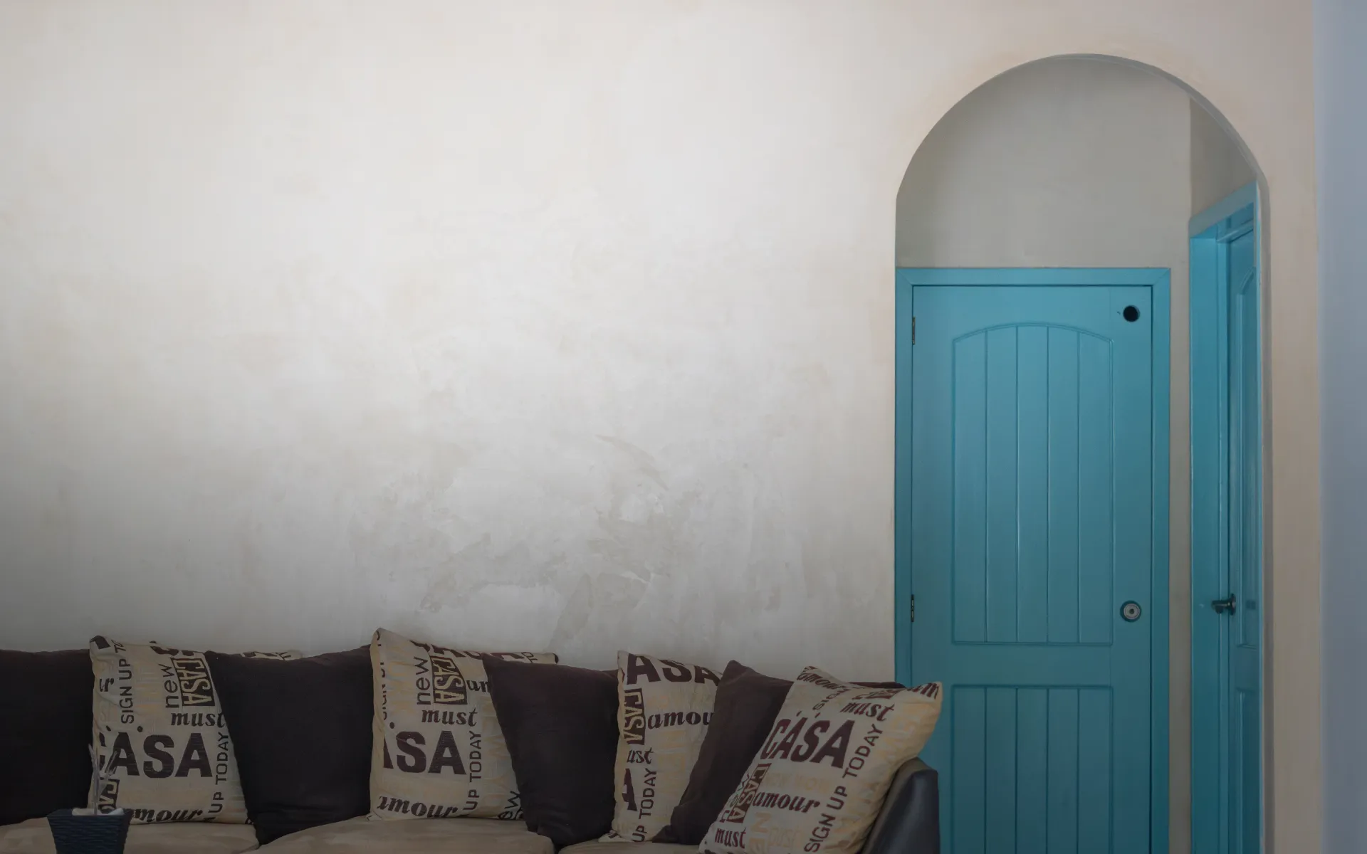 Pared blanca con un sofá negro y cojines estampados junto a una puerta arqueada azul.