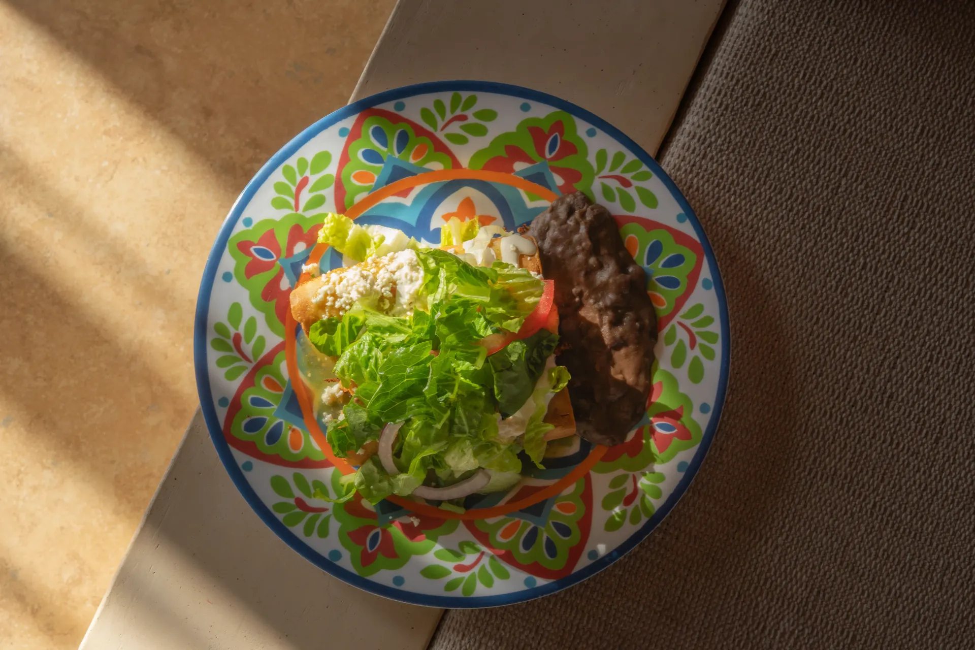 Plato colorido con ensalada y un plato oscuro a la parrilla sobre una mesa iluminada por el sol.