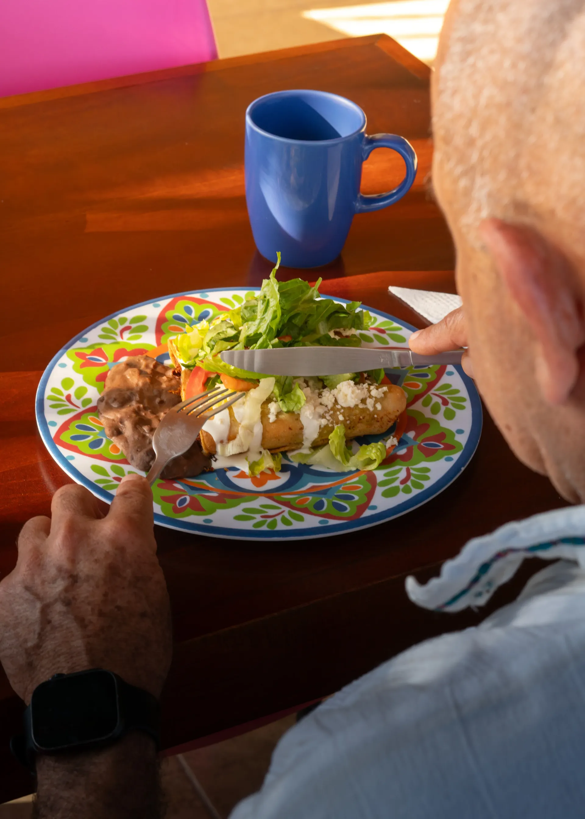 Una persona corta la comida en un plato colorido con una taza azul sobre una mesa.