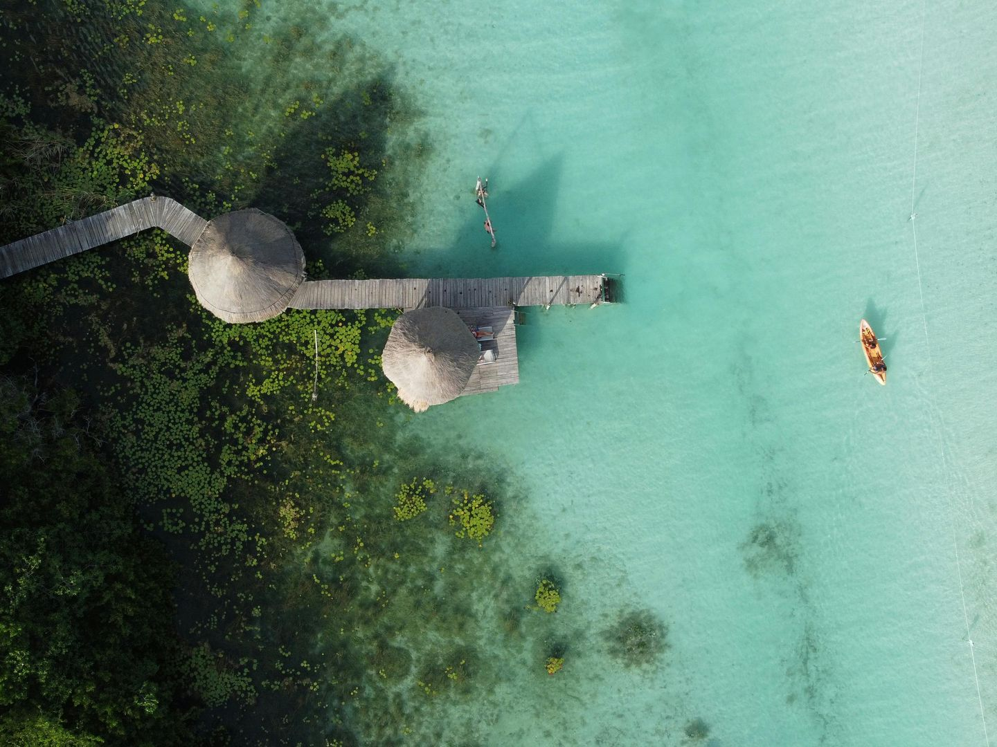 Vista aérea de un muelle que se adentra en aguas turquesas, con dos nadadores cerca y un pequeño bote a la derecha.