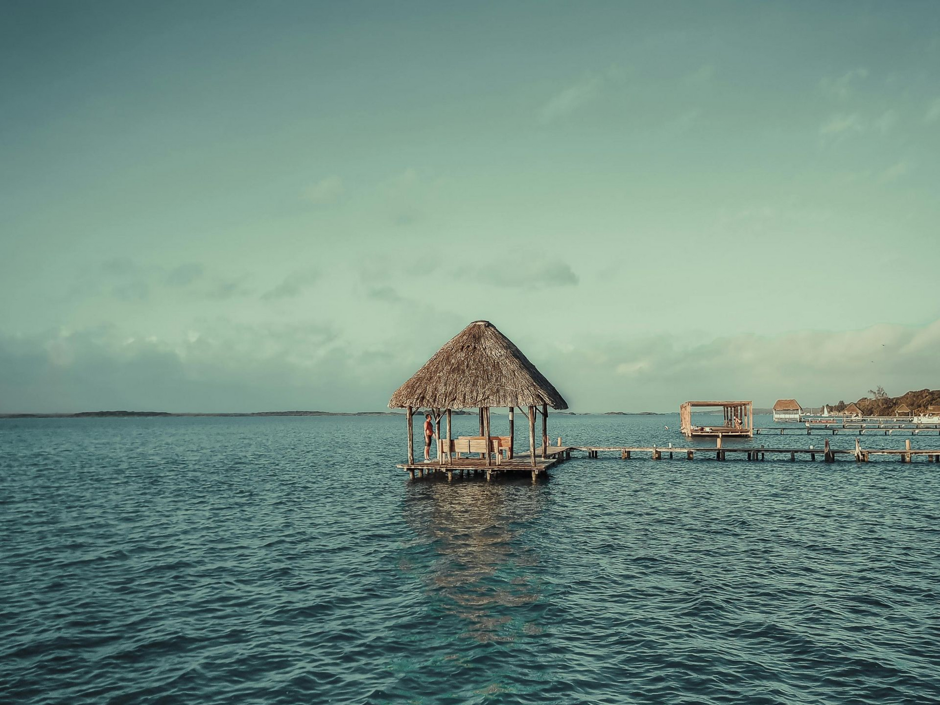 Cabaña con techo de paja sobre pilotes en aguas azules y tranquilas, con un muelle y pequeñas embarcaciones.