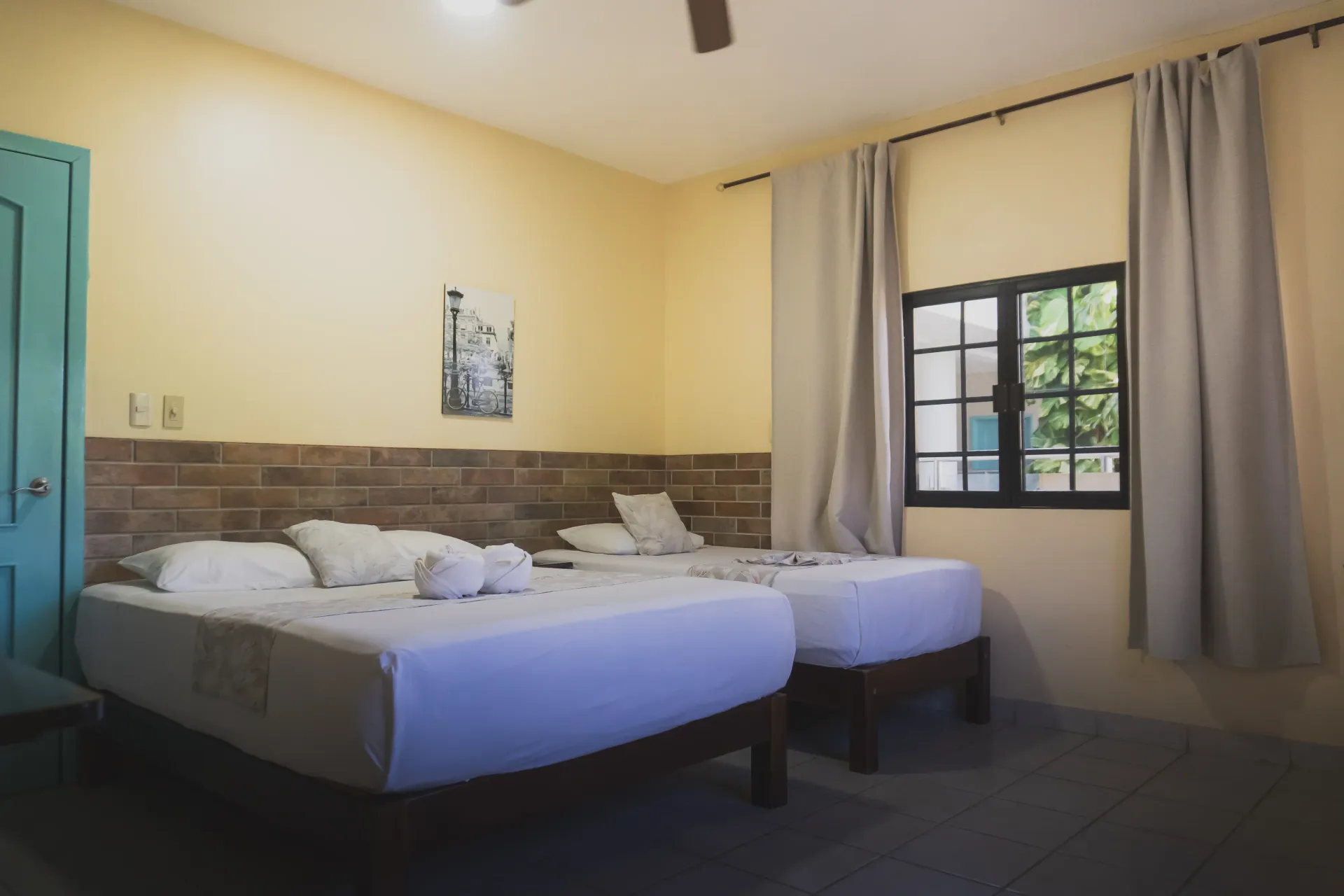 Habitación con dos camas individuales, sábanas blancas, paredes beige y una ventana con cortinas y vistas al exterior.