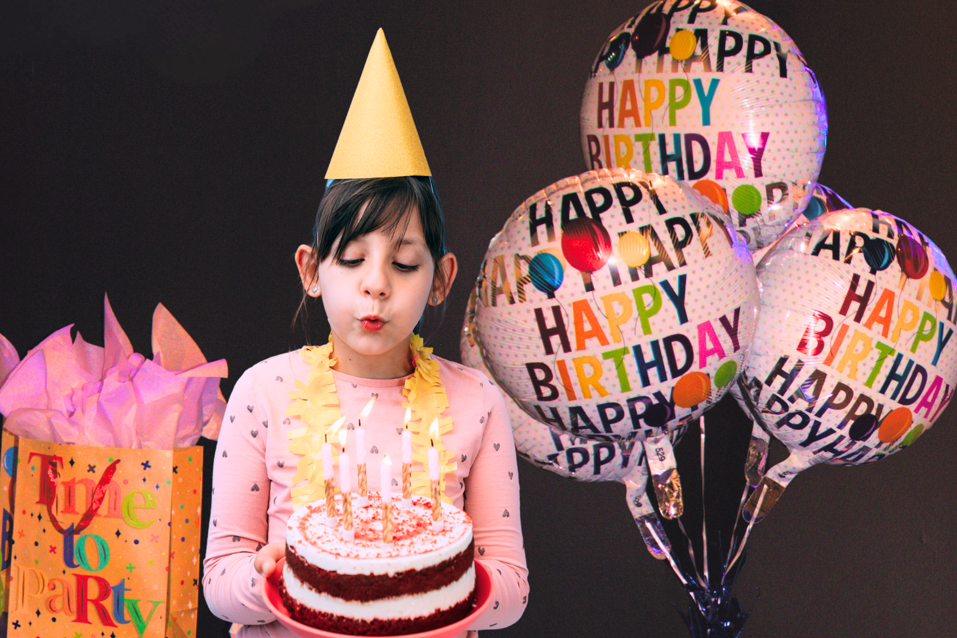 A young girl blowing out the candles on a birthday cake, adjacent to a bouquet of helium balloons.