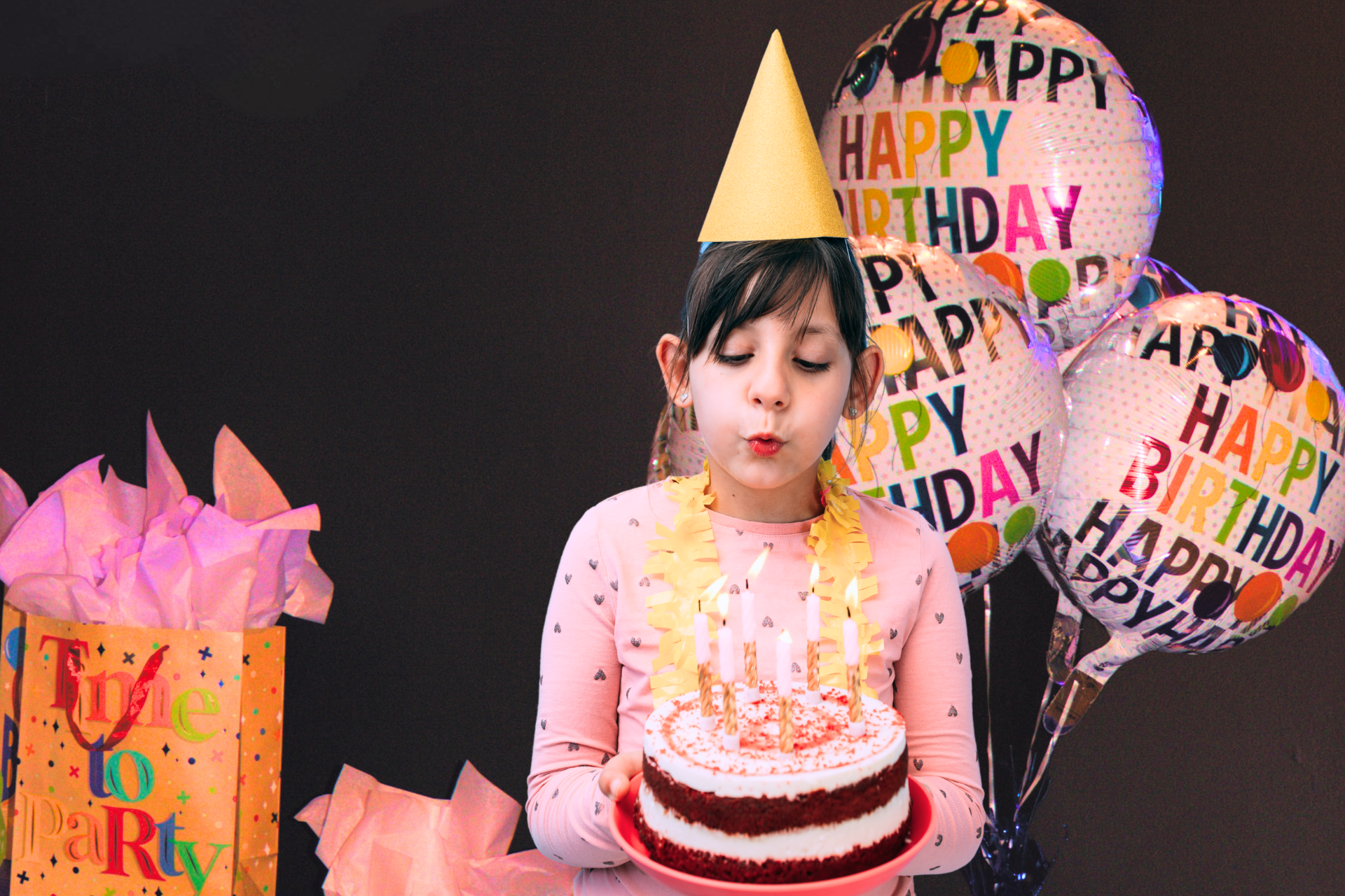 A young girls blowing out candles on a birthday cake in front of a bouquet of helium birthday balloons.