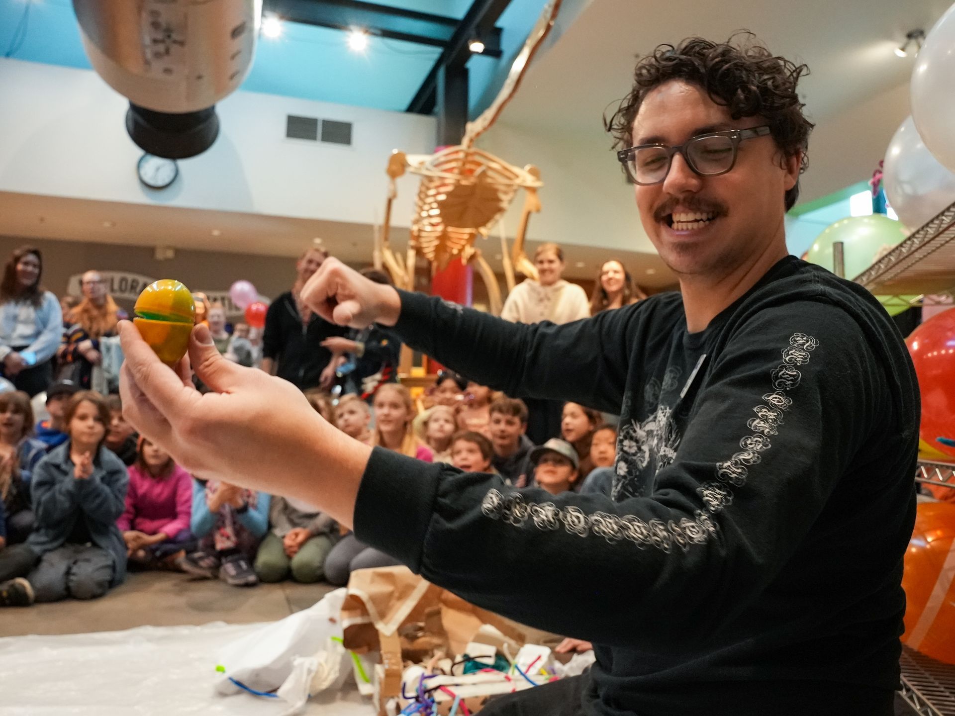 Man holding a plastic egg with paint coming out of it, gesturing to a crowd of children with a skeleton in the background, indoors at ScienceWorks.
