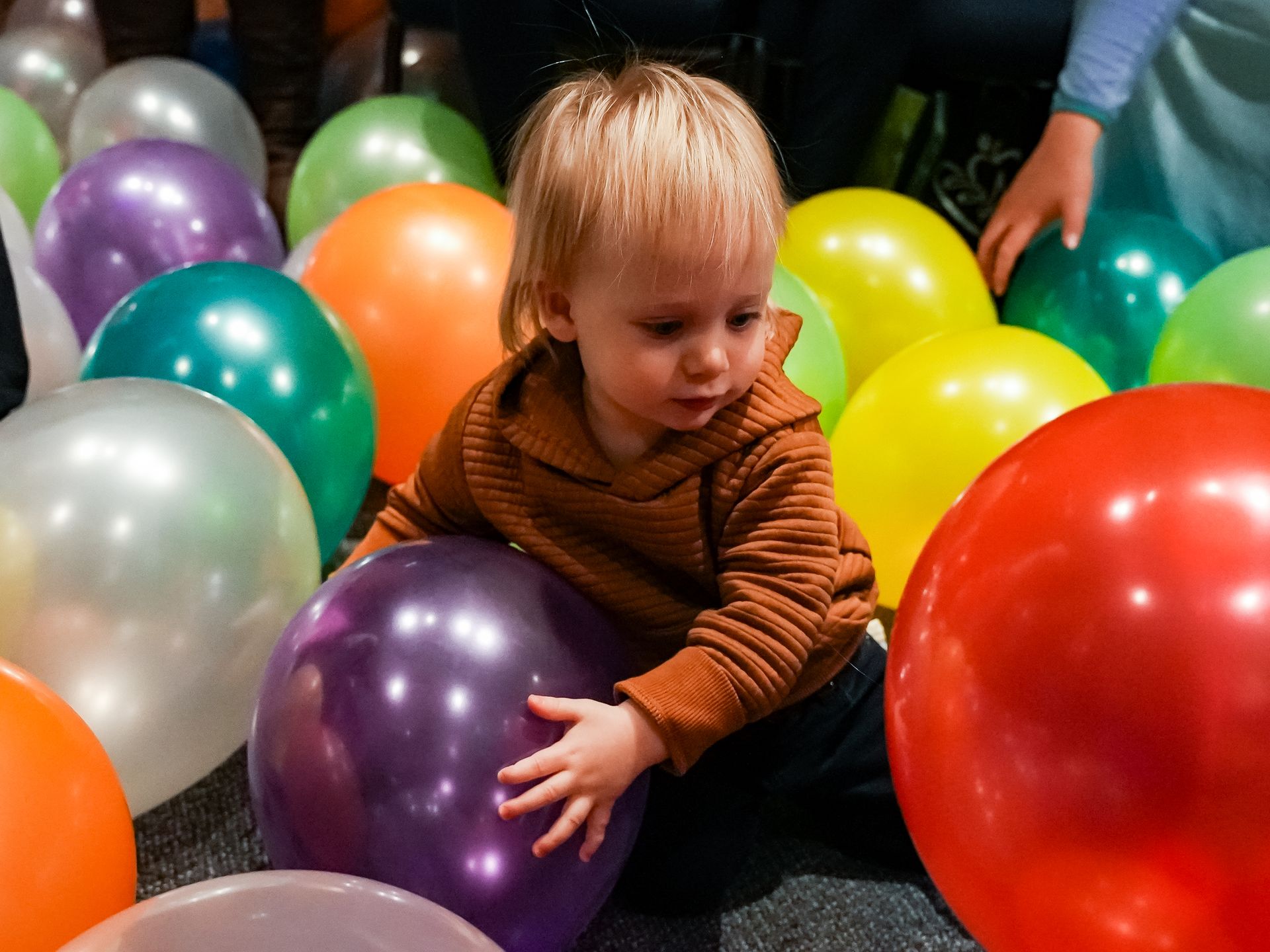 Young child in brown sweater surrounded by colorful balloons, touching a purple one at ScienceWorks.