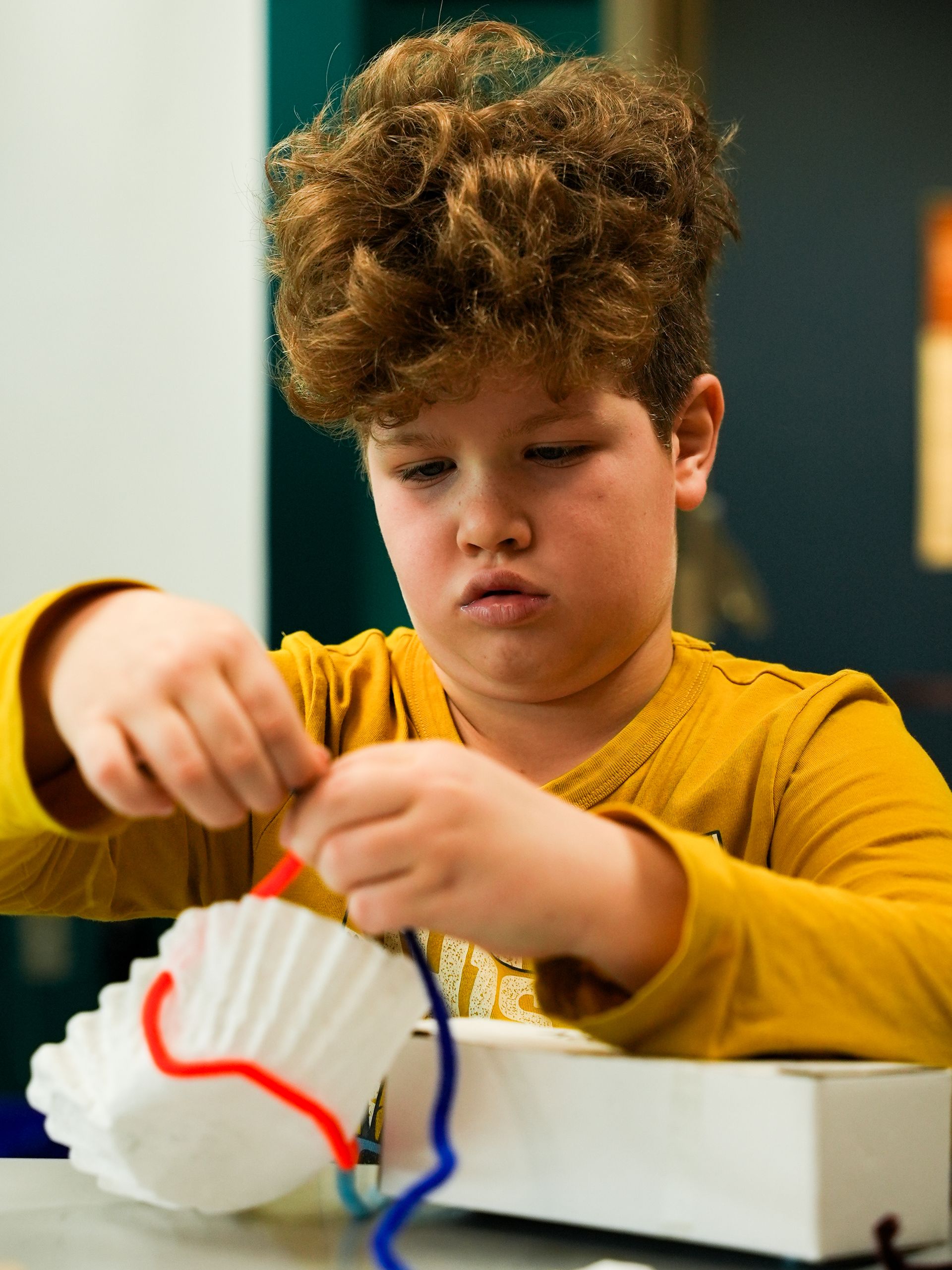 Boy with curly hair threading pipe cleaners through a coffee filter, focused expression at ScienceWorks.