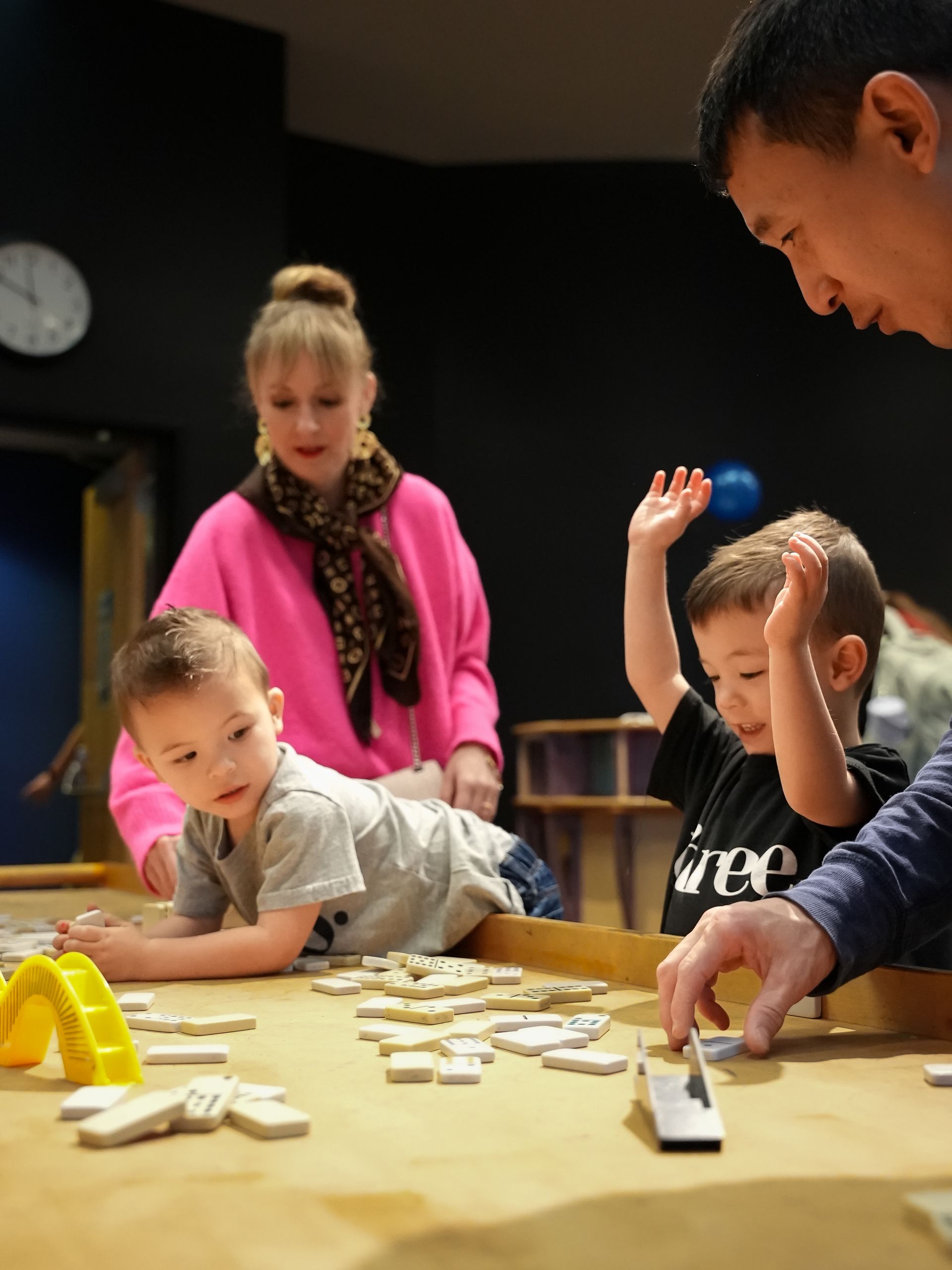 Children and adults play with dominoes at a table. One boy raises his arm. Others look on at ScienceWorks.