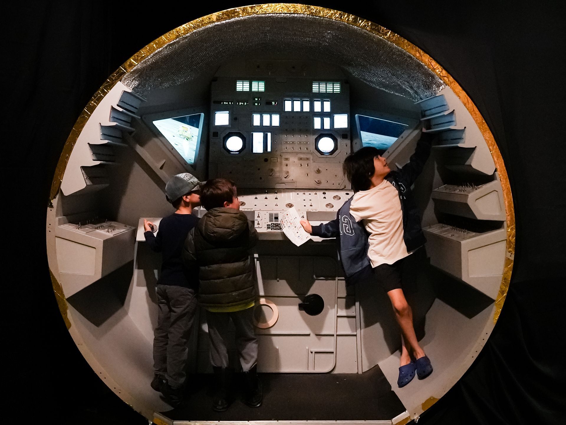 Children explore a model spacecraft interior, pointing at controls, within a museum exhibit at ScienceWorks.
