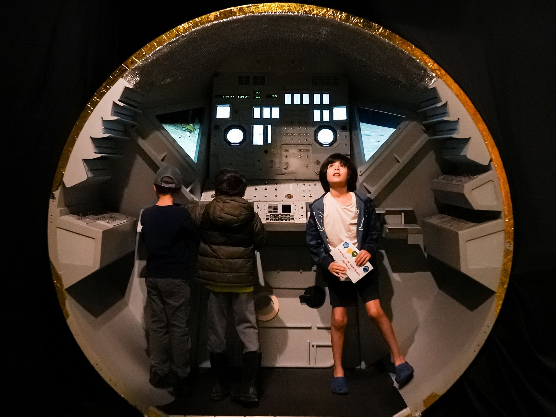 Children inside a spacecraft model at ScienceWorks, looking up at controls. Interior has a retro design, and gold trim.