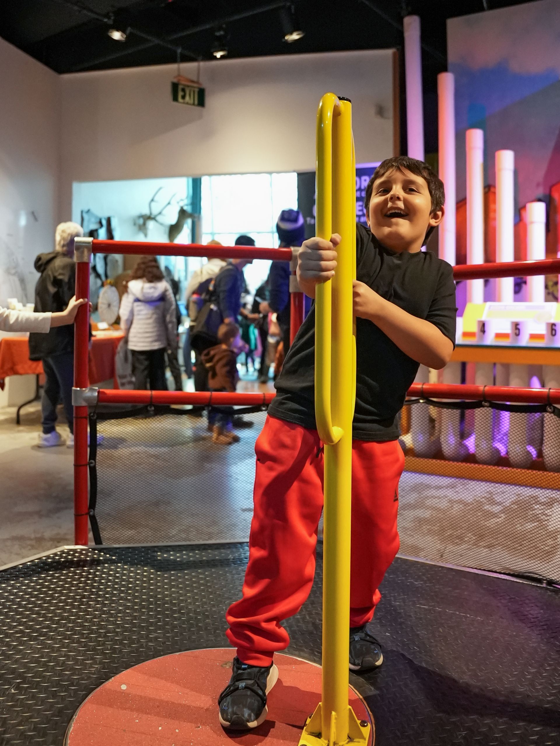 Boy holds yellow pole, smiling in a play area. Red pants, black shirt, and circular platform. Other people in background at ScienceWorks.