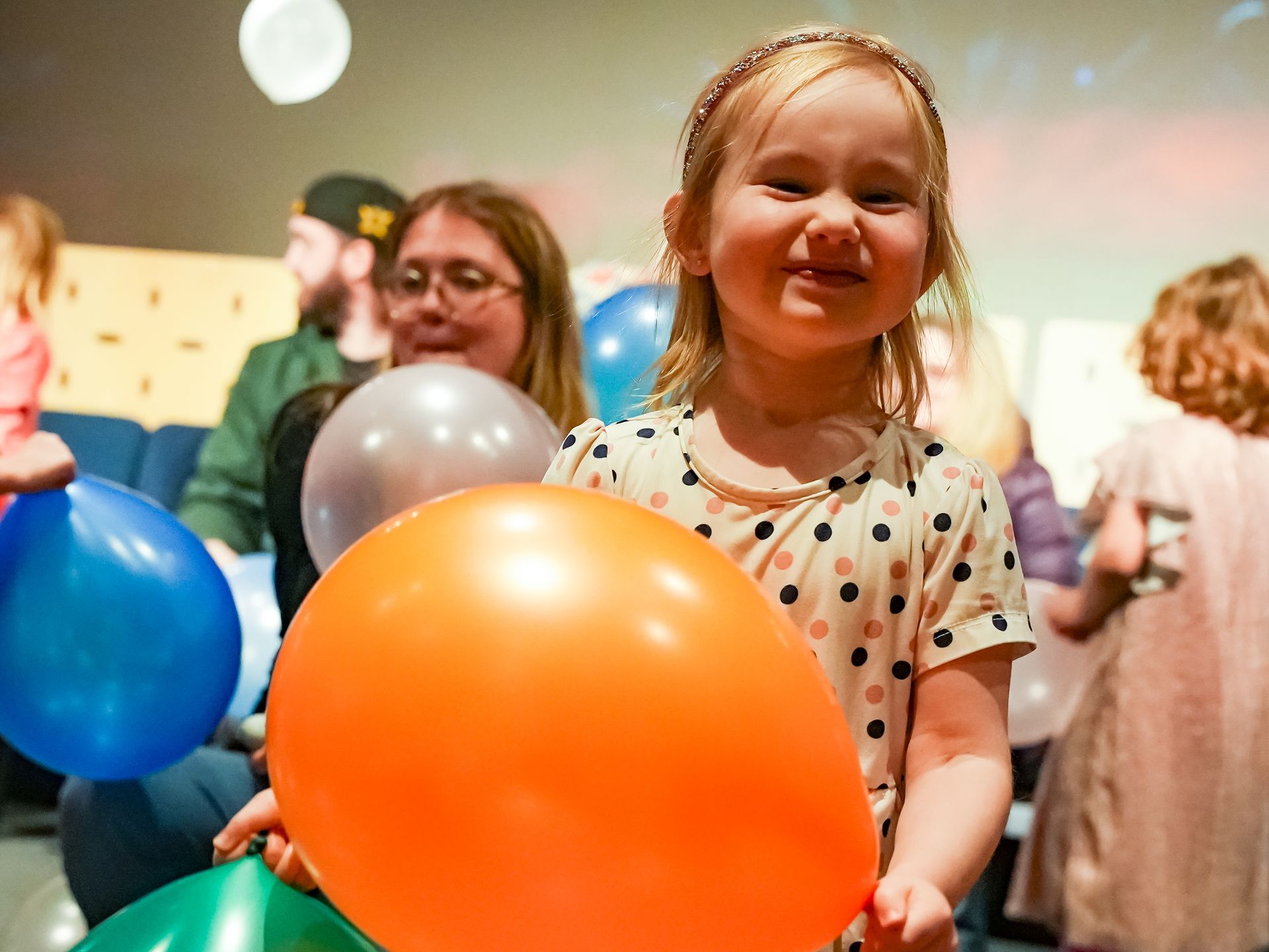Smiling child holding orange balloon; surrounded by other balloons and people indoors at ScienceWorks.