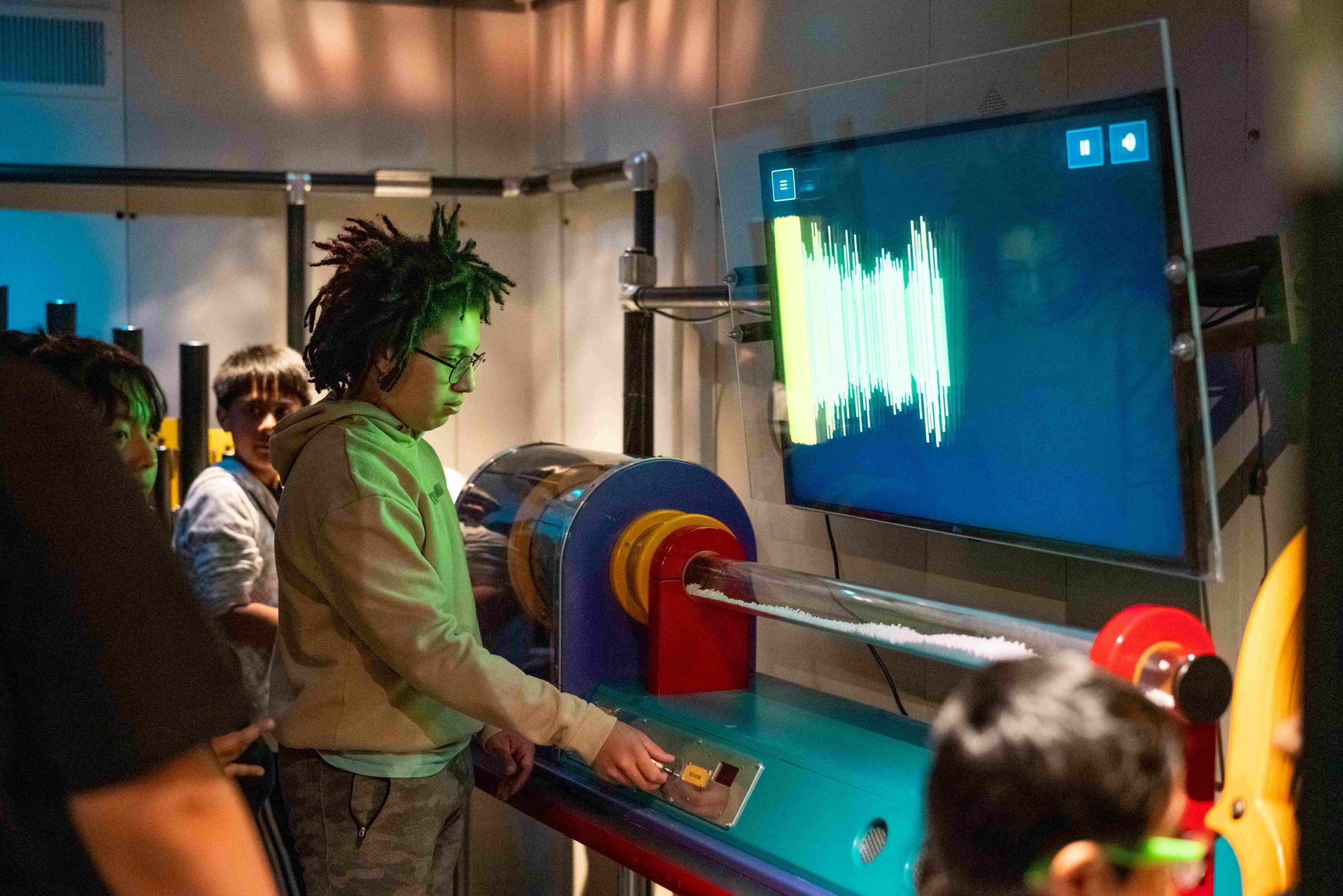 Boy with locks using a science exhibit at a museum, displaying a wave pattern on screen.