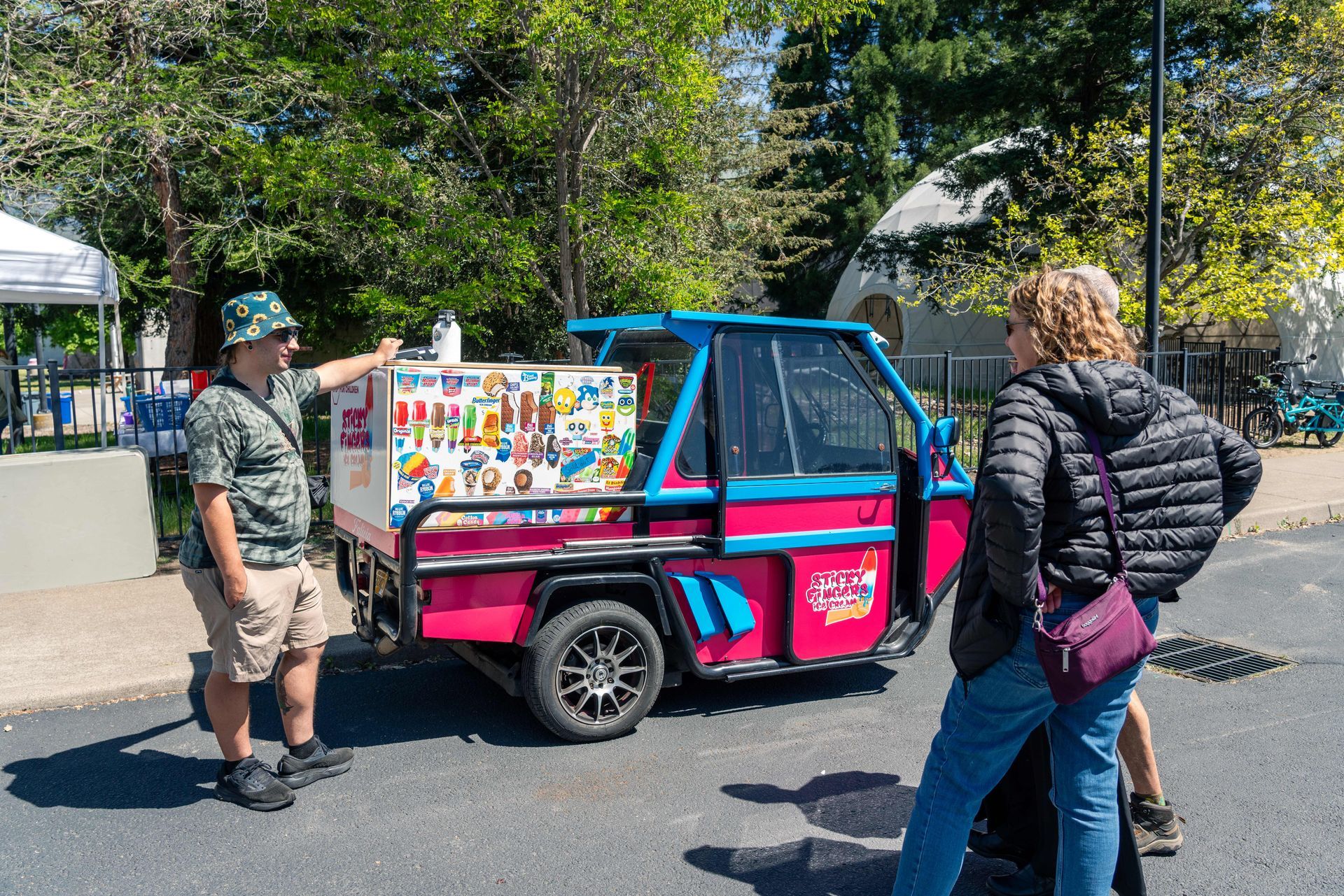 Two people stand by a pink-and-blue street cart with colorful menu signs outdoors.