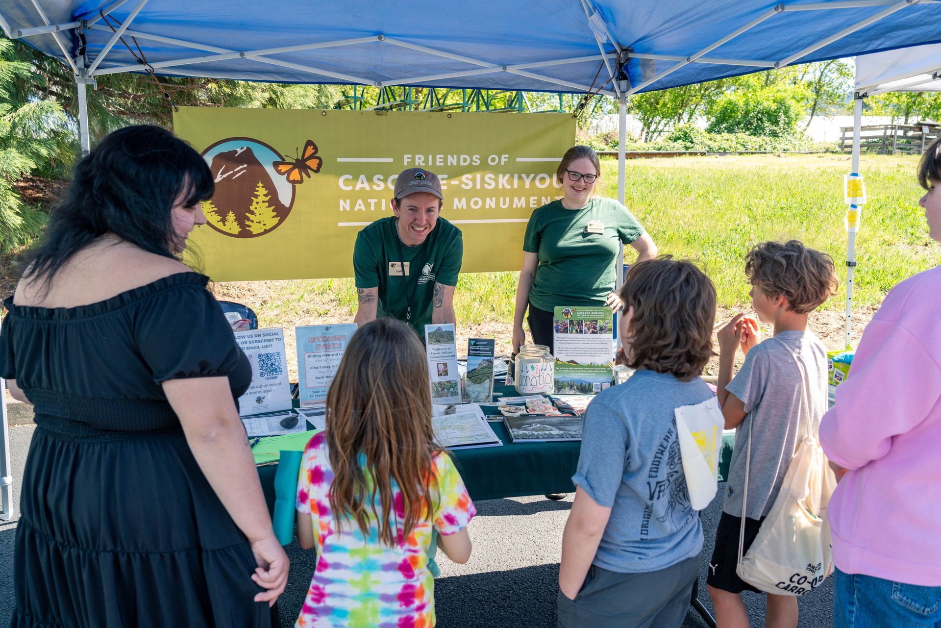 Two people at a community outreach booth under a blue tent, with children gathered around tables of materials.