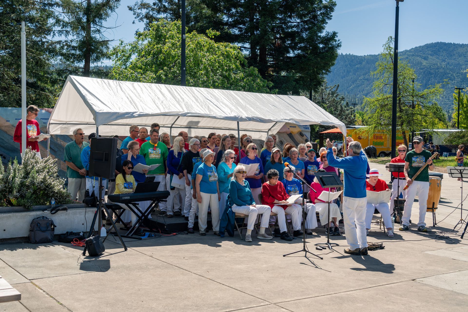 Outdoor band performing in blue shirts under a white canopy, with a seated audience nearby on a sunny day