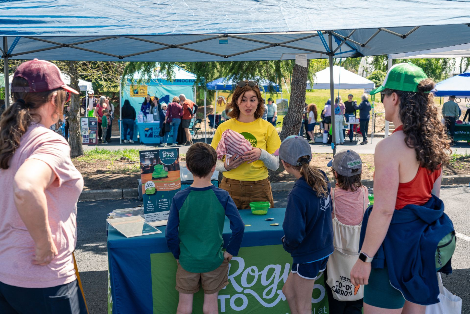 People at a community outreach booth under a white tent, with children gathered around tables of materials.