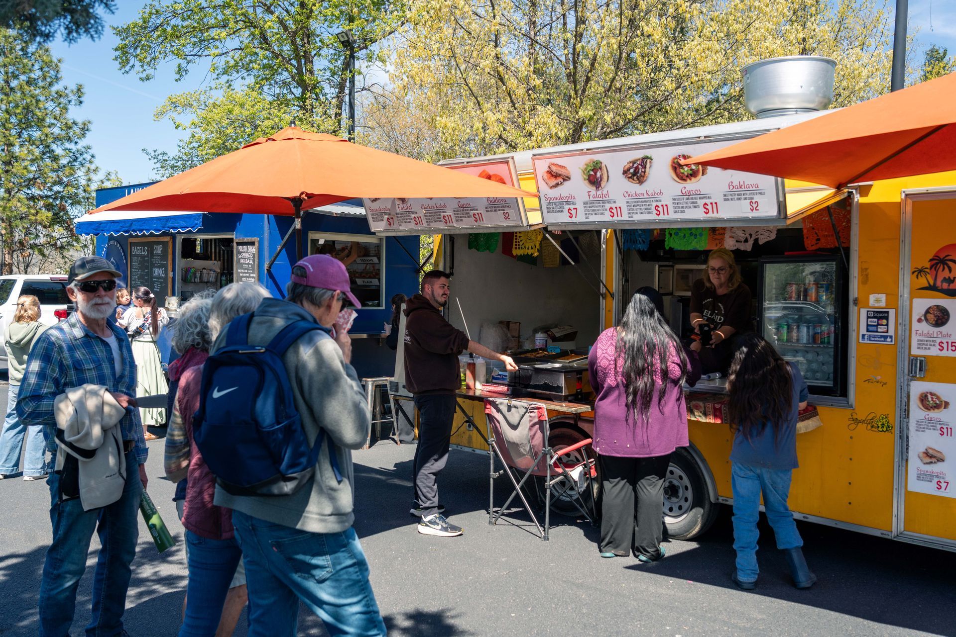 Outdoor food stand with orange umbrellas and people ordering from a colorful menu sign