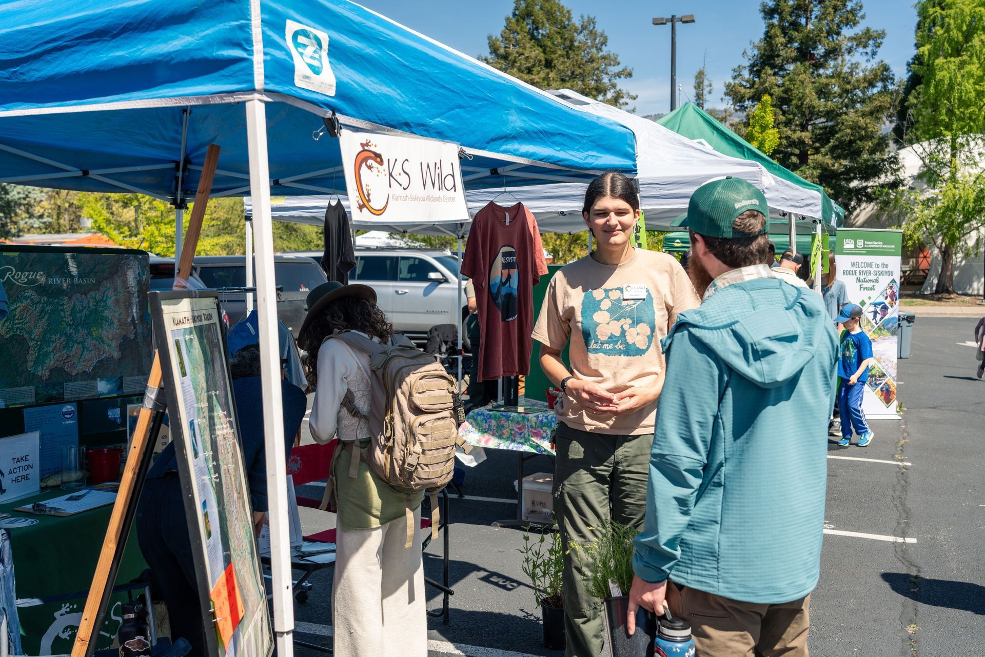 People chatting at an outdoor festival under blue canopies in a sunny parking lot