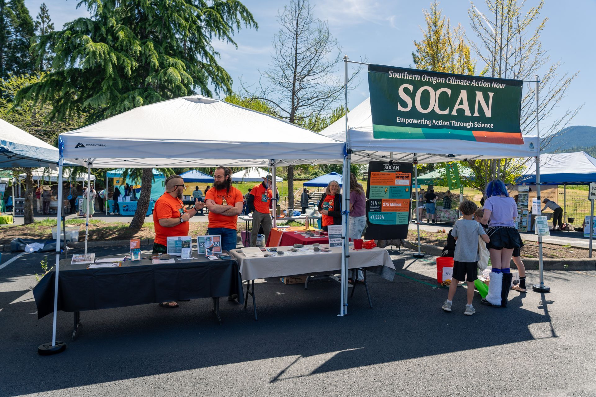 People at a community outreach booth under a white tent, with children gathered around tables of materials.