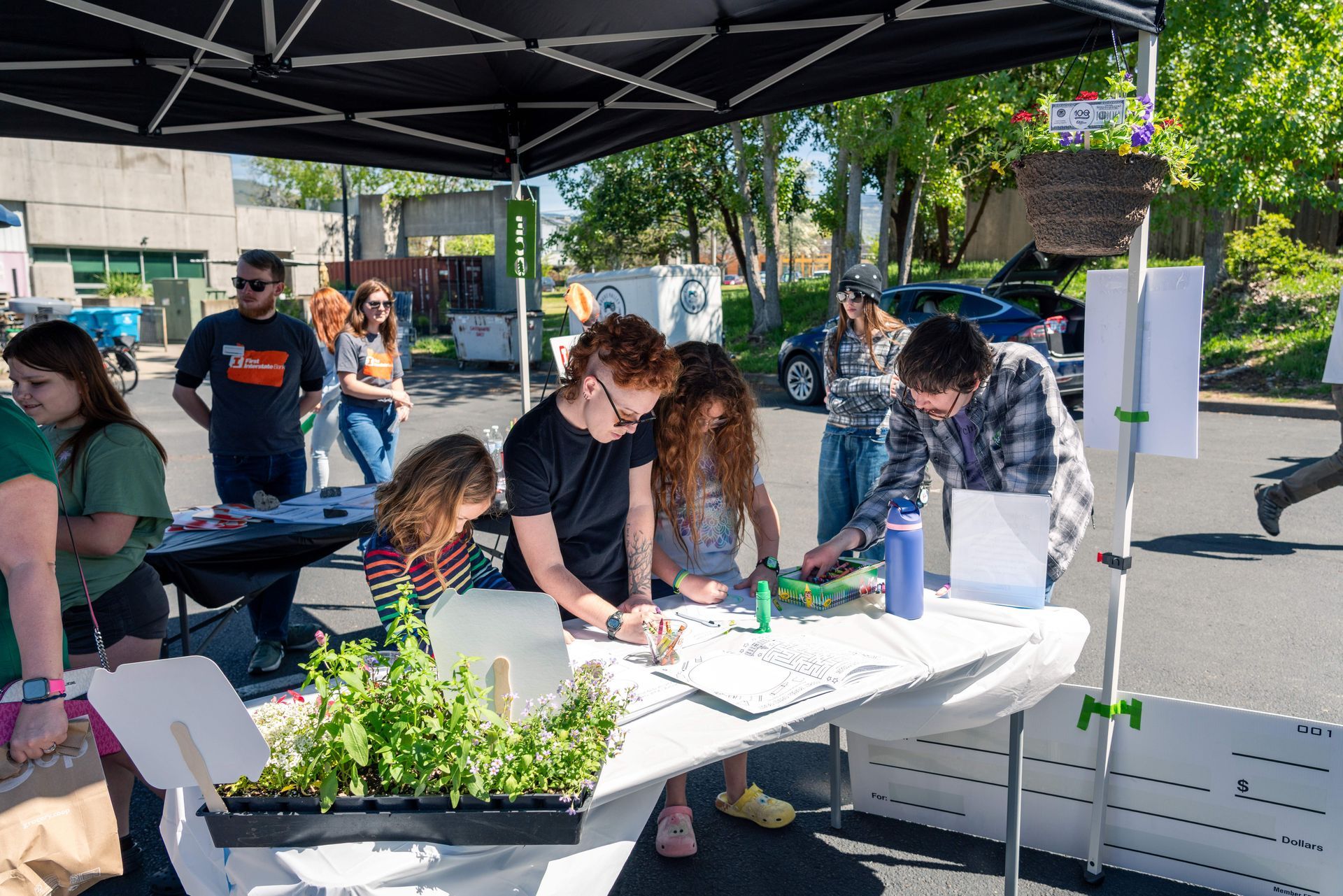 People drawing at an outdoor community booth under a canopy, with tables and potted plants.