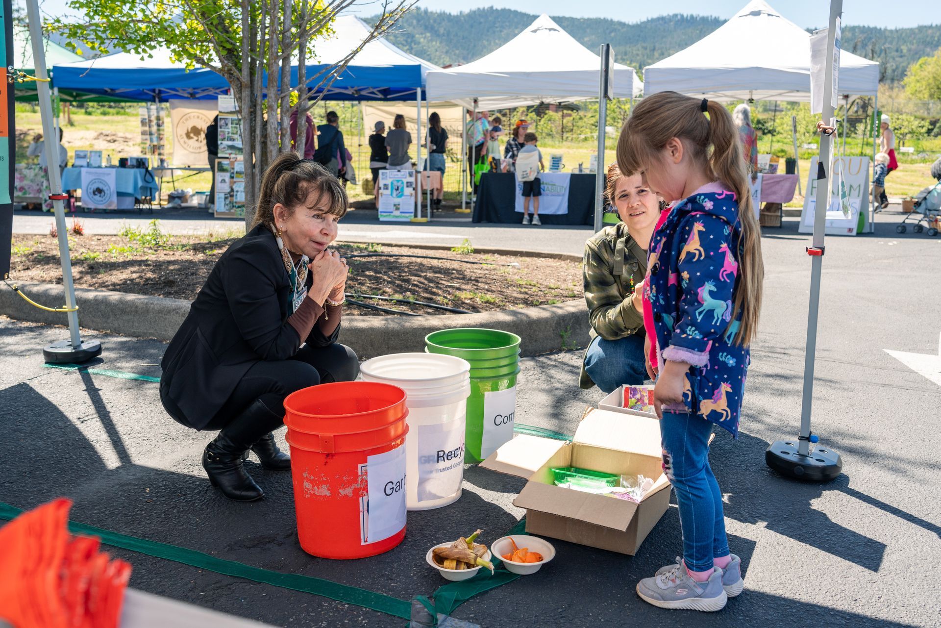 Woman crouches with child at outdoor market, sorting food and other waste into bright buckets and boxes.