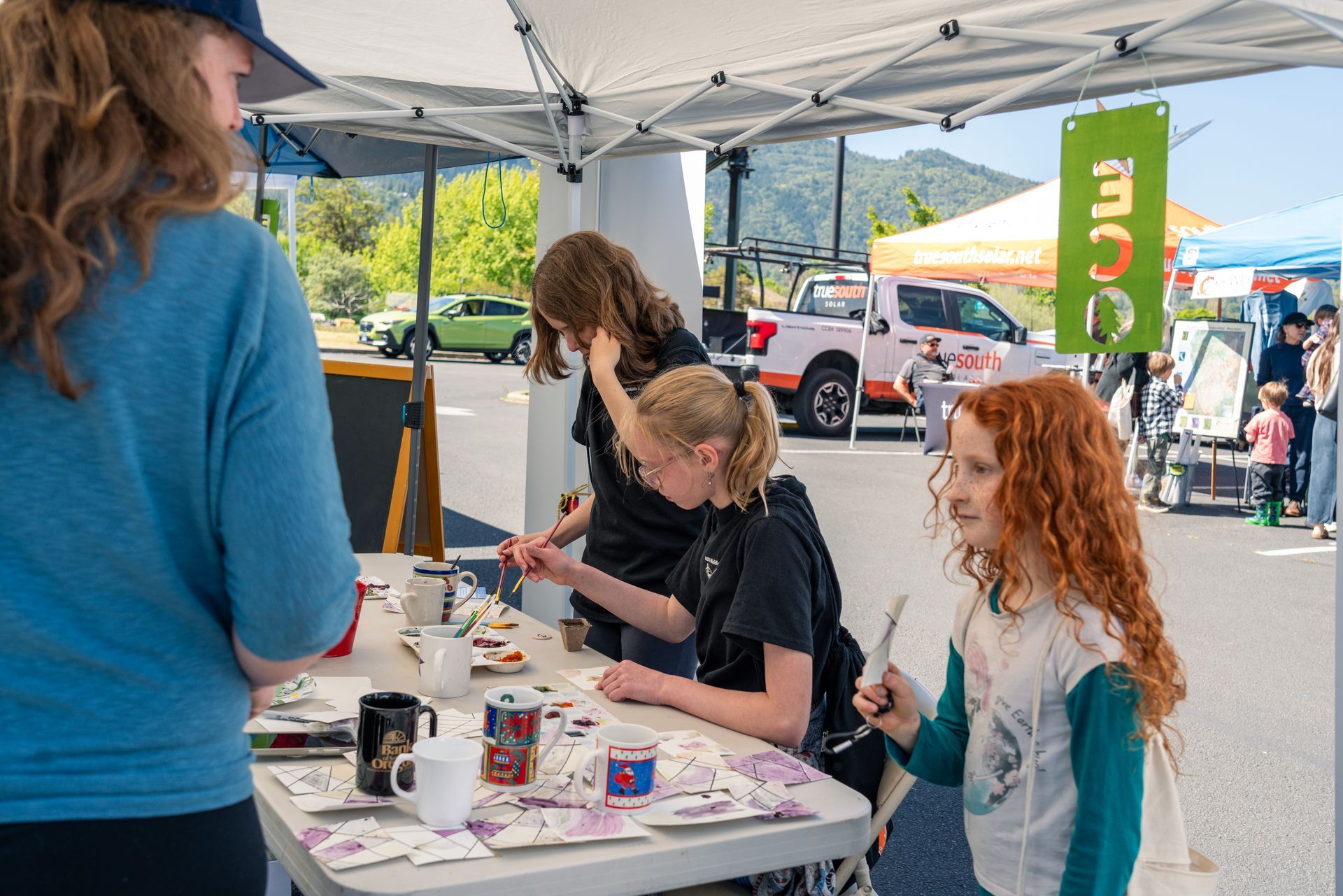 People at a community outreach booth under a white tent, with children gathered around tables of materials.