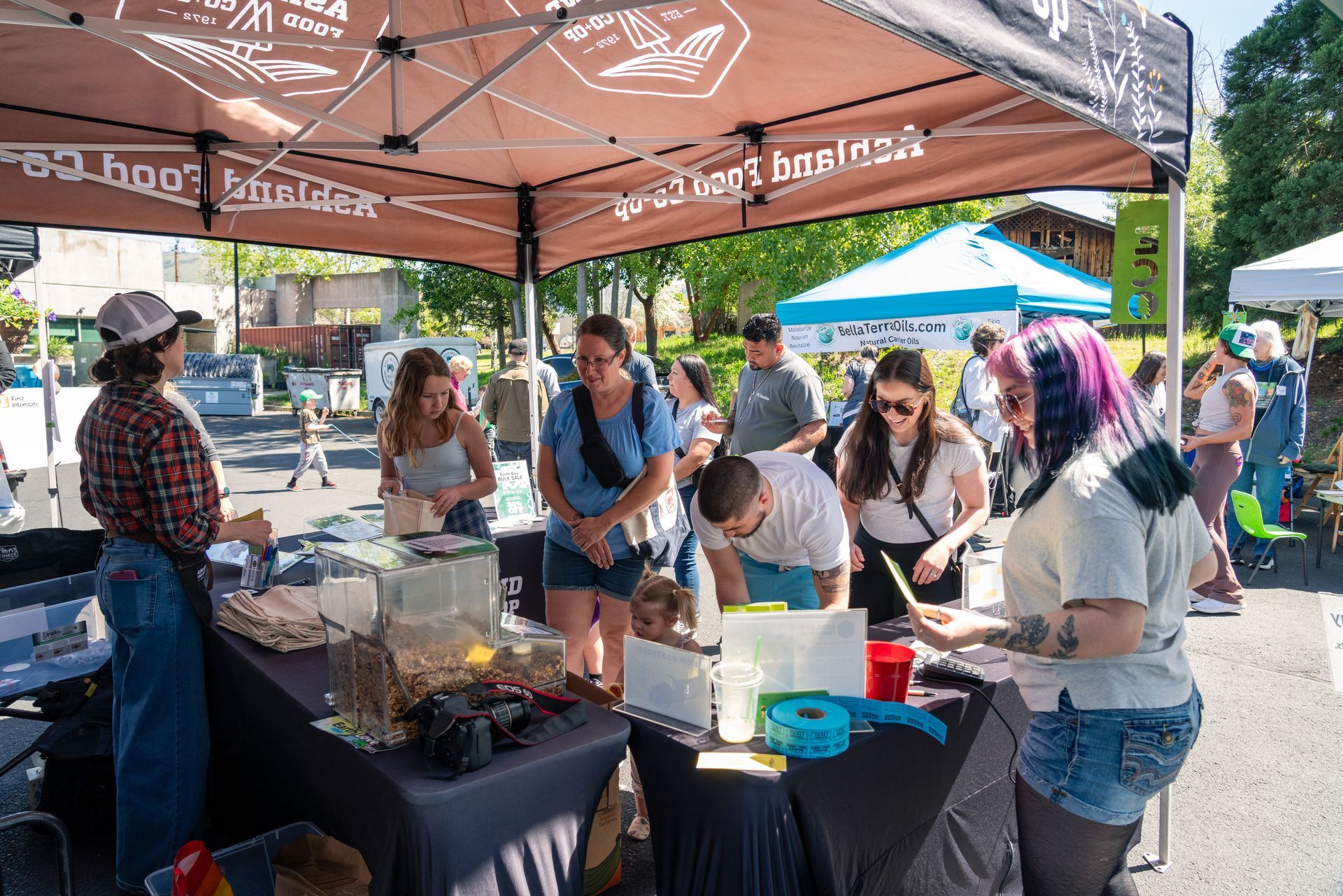People browsing and sampling food at an outdoor event under a brown canopy