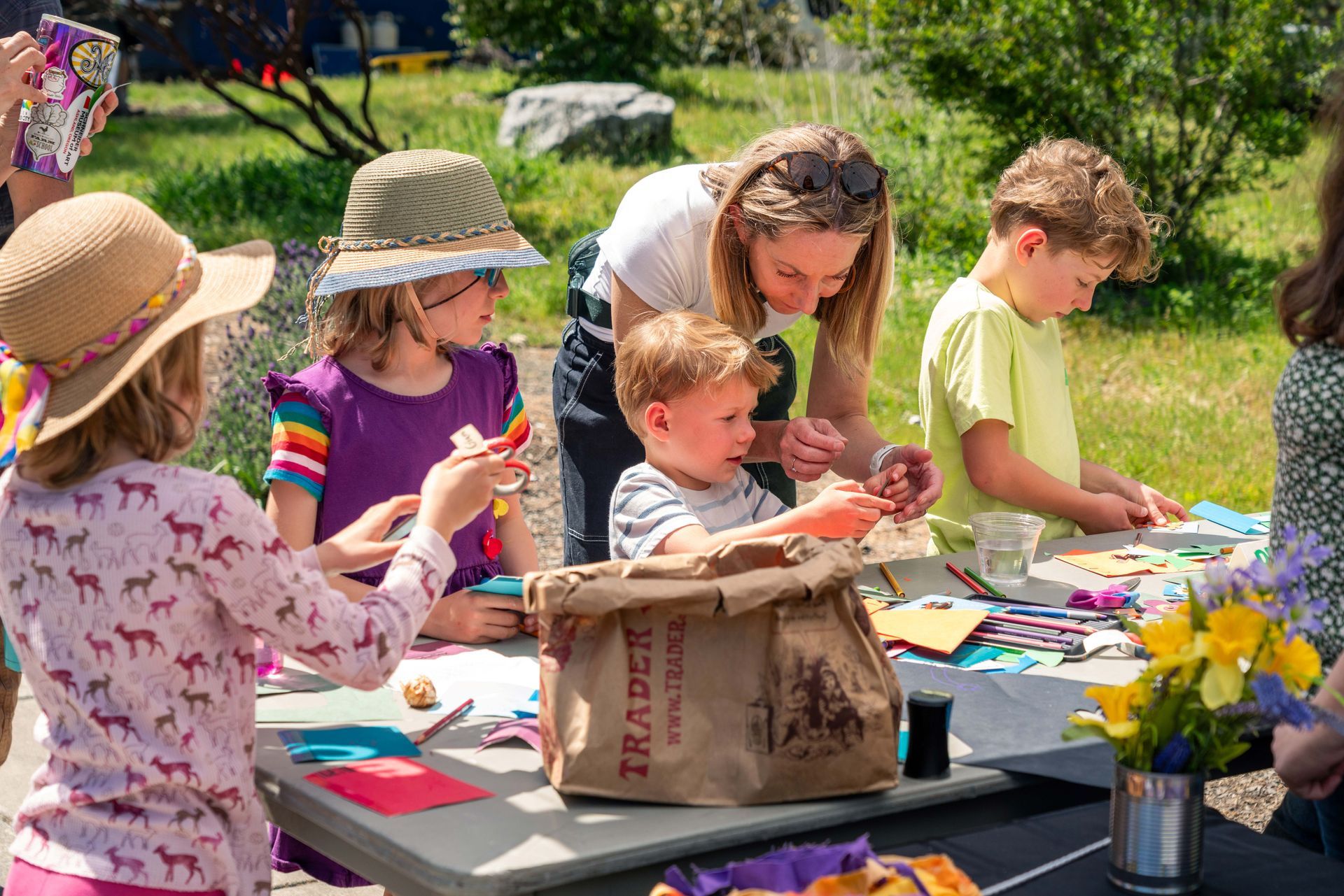 Children making crafts at a table outdoors, with an adult helping and supplies spread across the table.