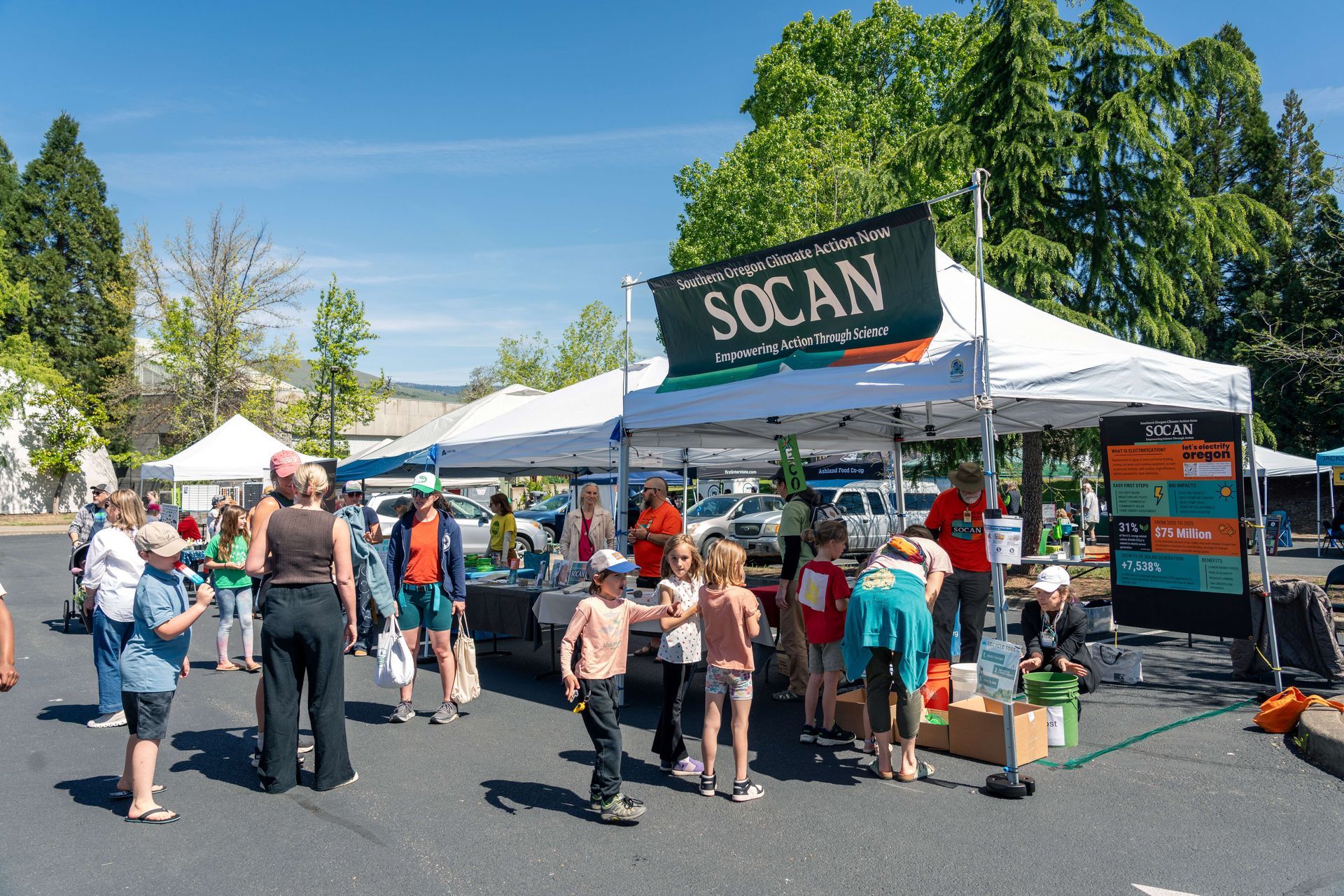 People at a community outreach booth under a white tent, with children gathered around tables of materials.