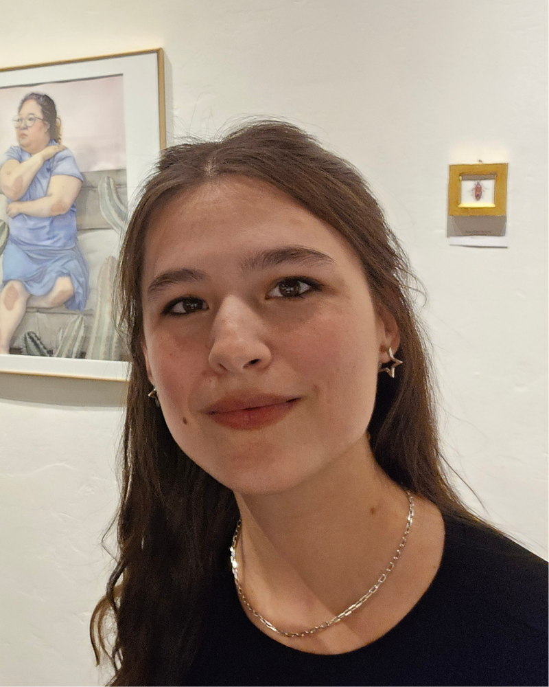 Photo of the artist Kyrianna smiling at the camera, art on white wall behind.