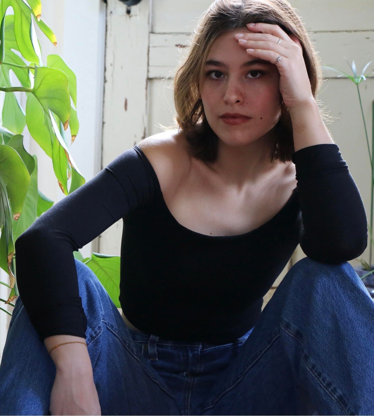 Woman in black top and jeans, hand on forehead, seated by plants and a door. It is the artist KYRIANNA.