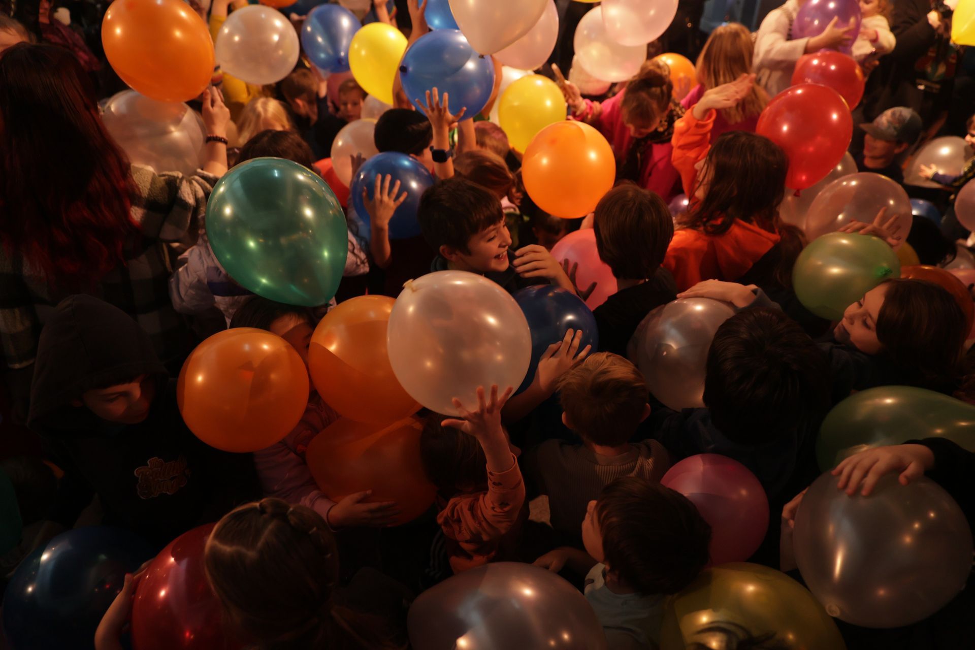 People in a crowd surrounded by colorful balloons, raising arms at ScienceWorks.