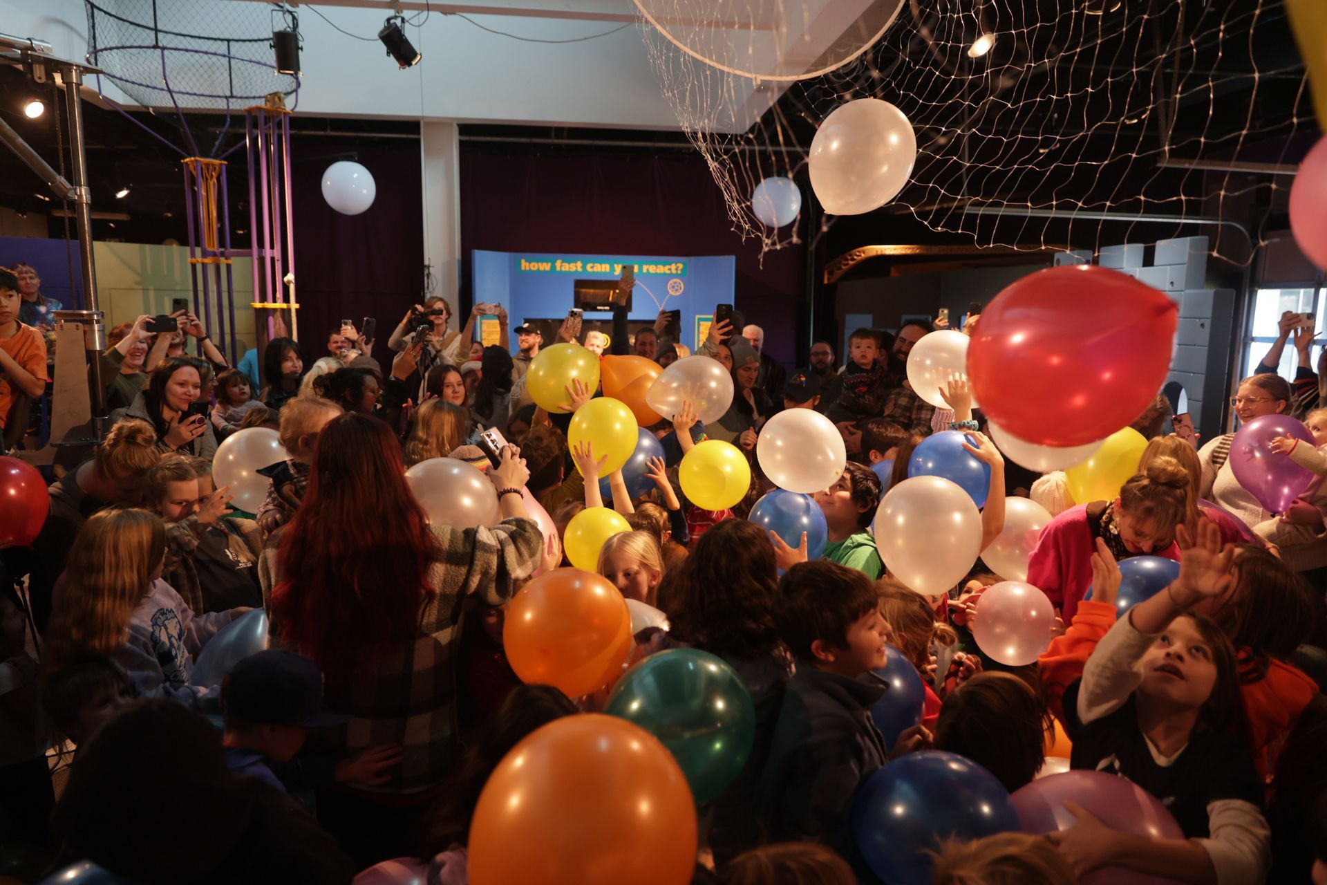 People releasing colorful balloons in a crowded room. Children and adults reach for balloons; festive occasion at ScienceWorks.