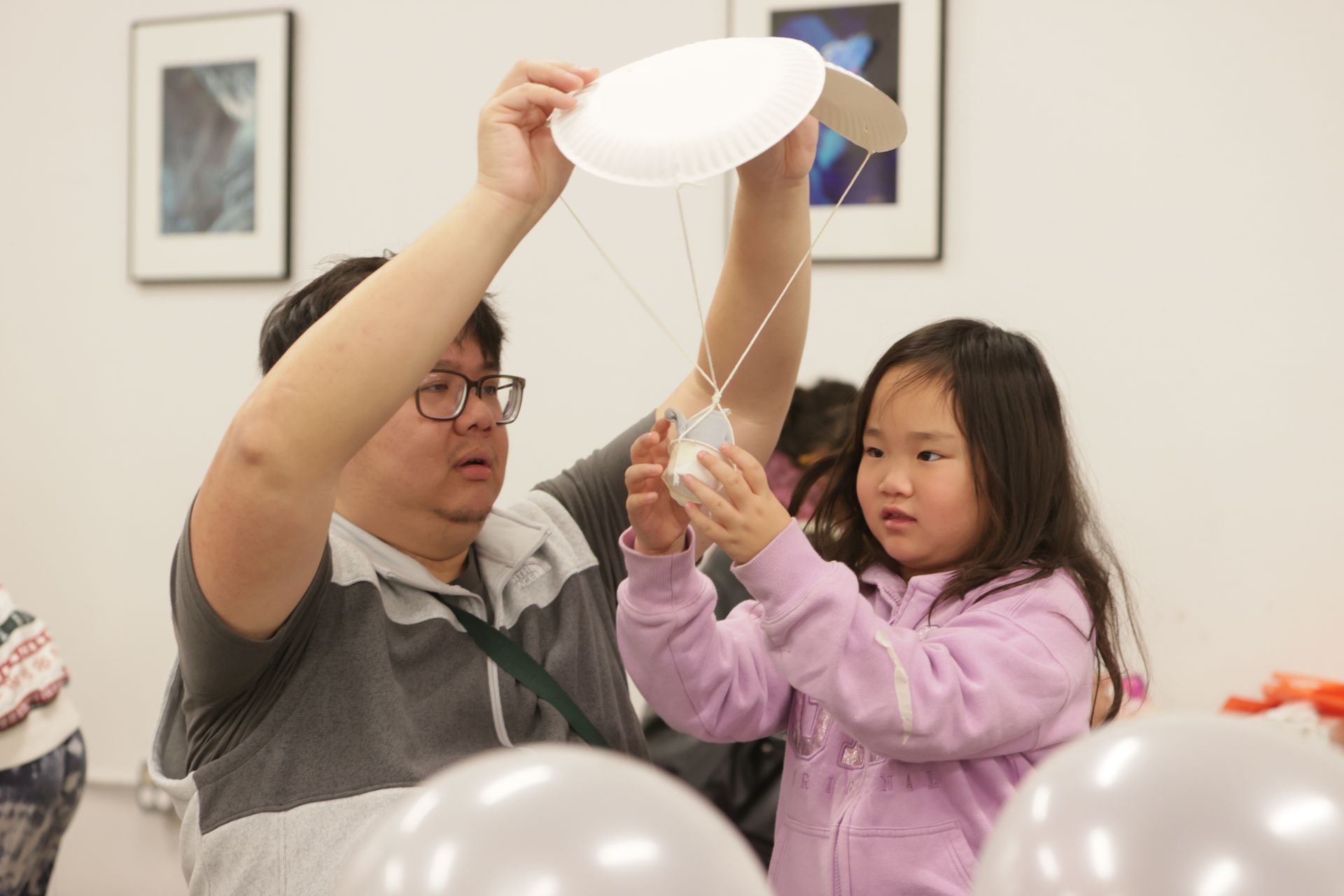Man and child building an egg drop craft with a parachute together, indoors at ScienceWorks. The man holds the plate above.