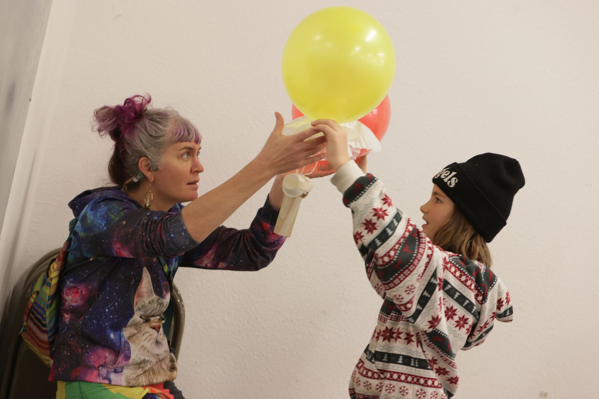 Woman and child holding their egg drop contraption made of cardboard and balloons  at ScienceWorks. The woman in a galaxy sweatshirt, child in a sweater and beanie. White wall background.