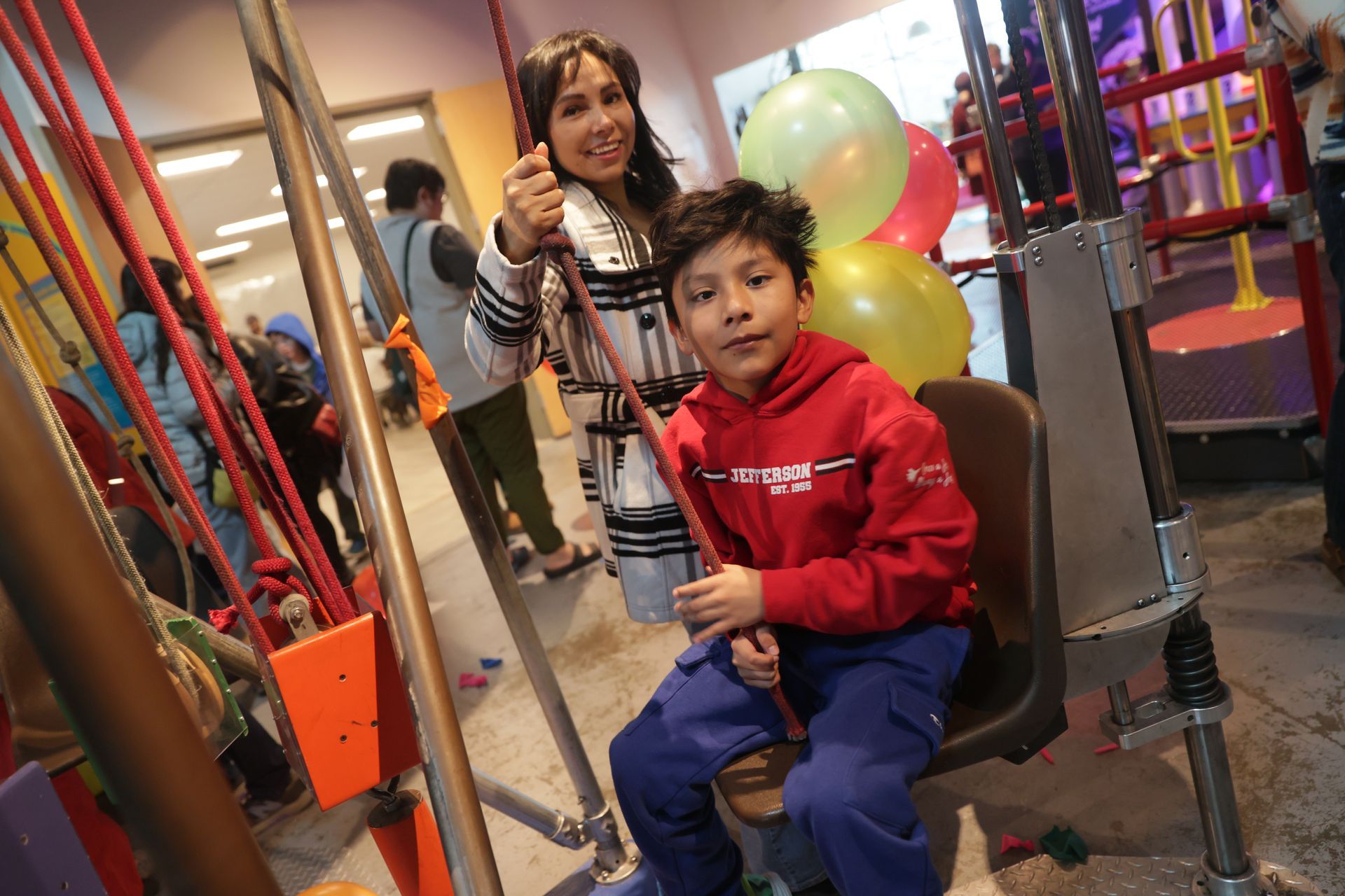 Woman, and a child in a red hoodie on a pulley chair at ScienceWorks. They both hold a rope. Balloons in the background.