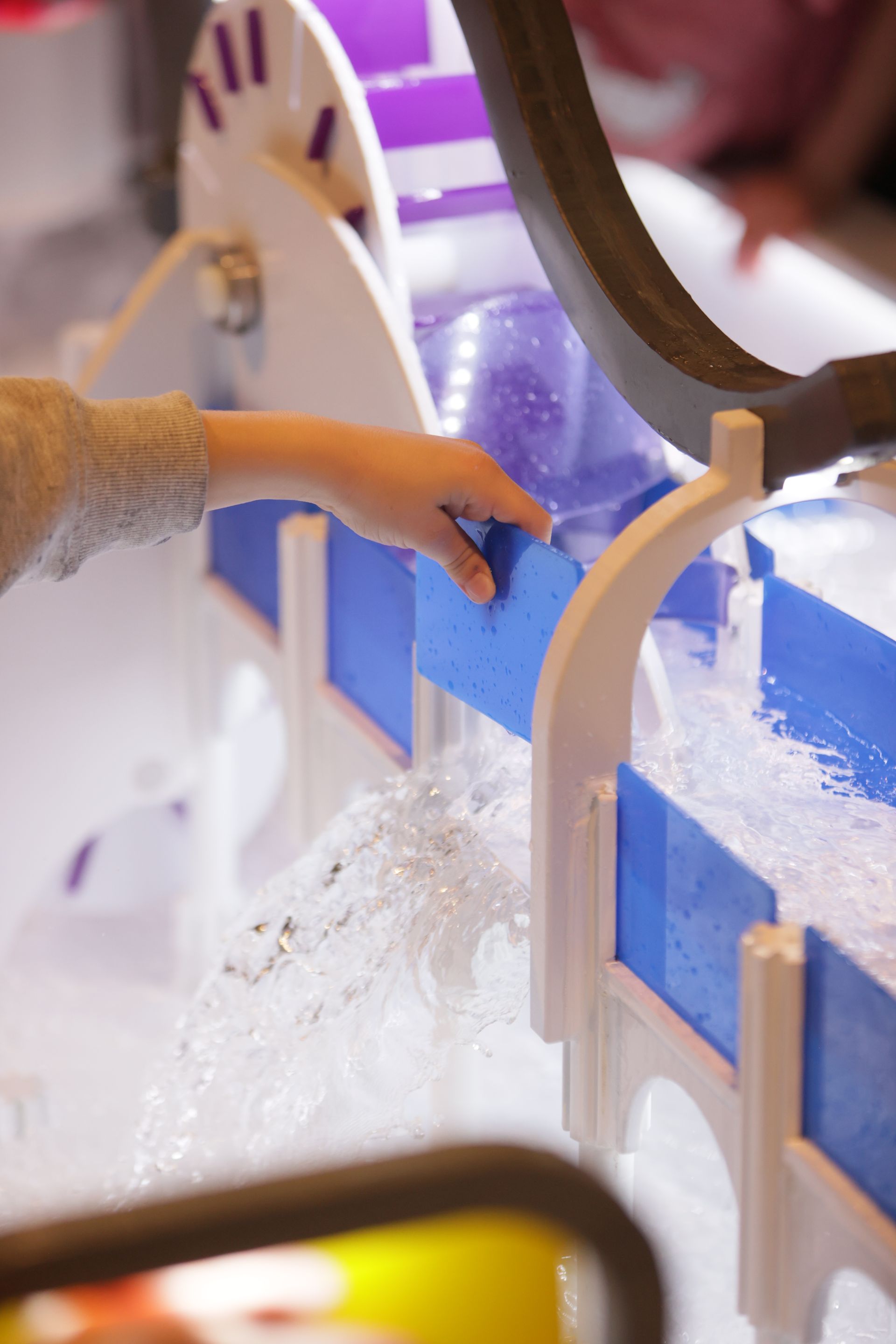 Child's hand interacting with a water flow exhibit at ScienceWorks. White and blue structure with purple accents. Water cascading.