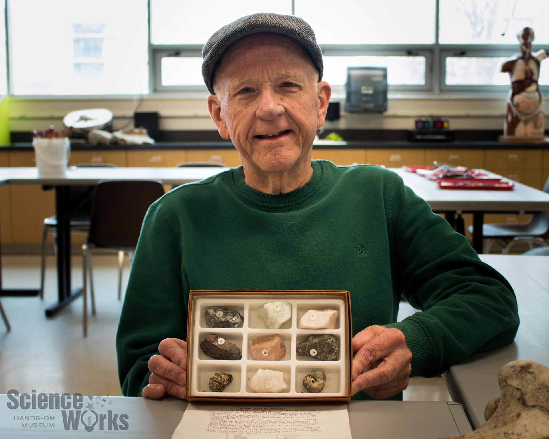 An elderly volunteer smiles at the camera and holds up tray of large fossils and minerals
