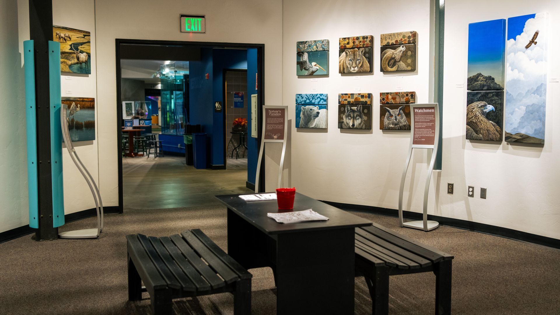 An entryway with a small table and benches. Artwork and information placards adorn the walls. Claire Duncan exhibit.