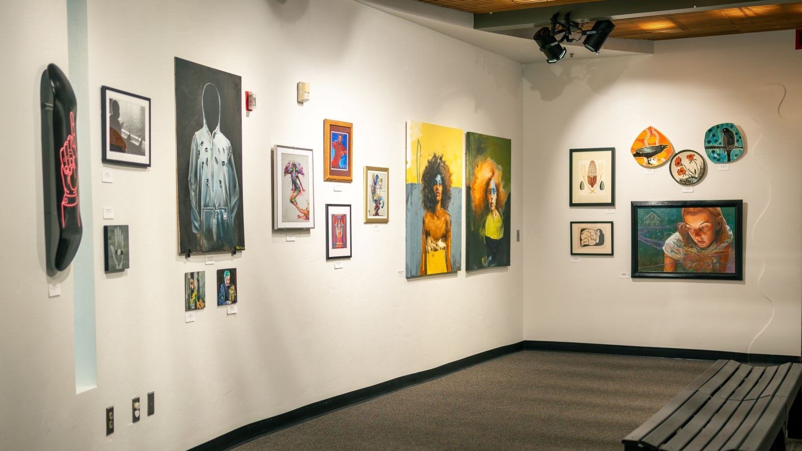 Art gallery interior with paintings on white walls; a bench is in the corner. Mural Fest 2025 exhibit.